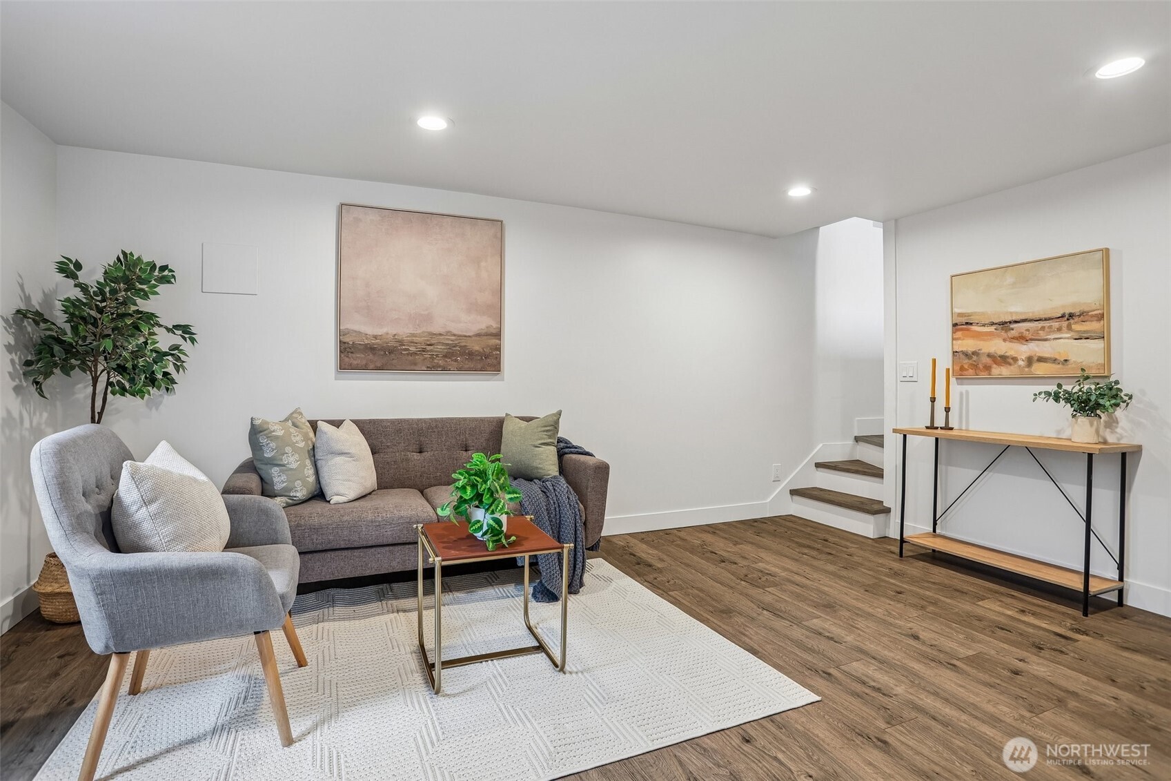 Staged living room with a gray sofa, armchair, white rug, and stairs.