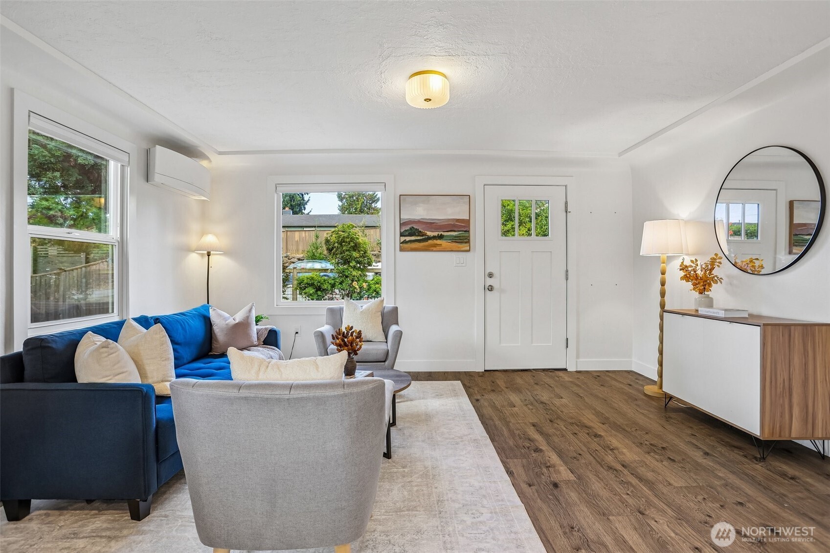 Staged living room with a blue sectional, gray chairs, wood floors, and large windows.