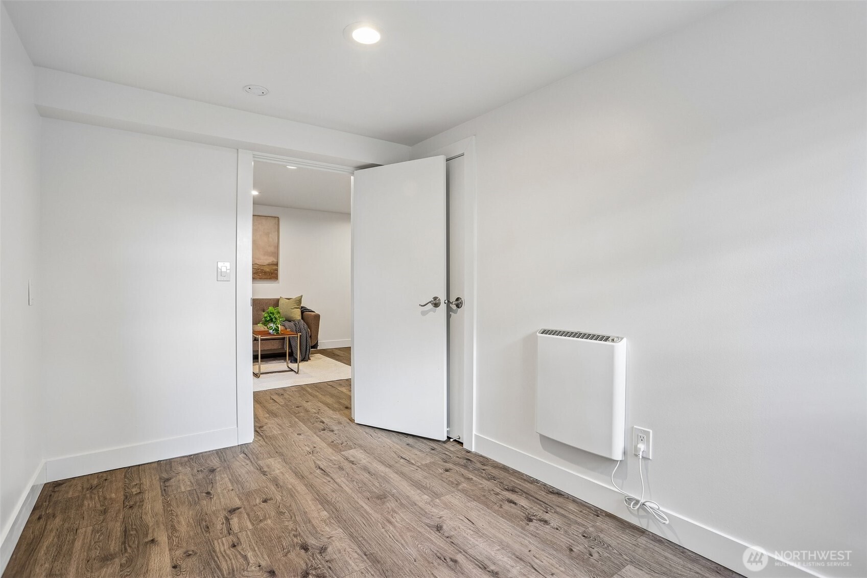 Empty room with wood-look flooring and a wall heater, looking through an open door to a furnished sitting area.