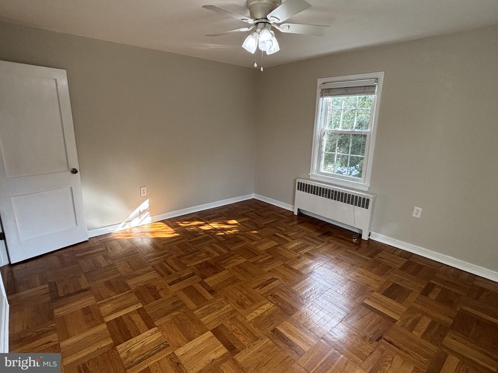 Empty bedroom with patterned parquet flooring, a ceiling fan, and a window above a radiator.