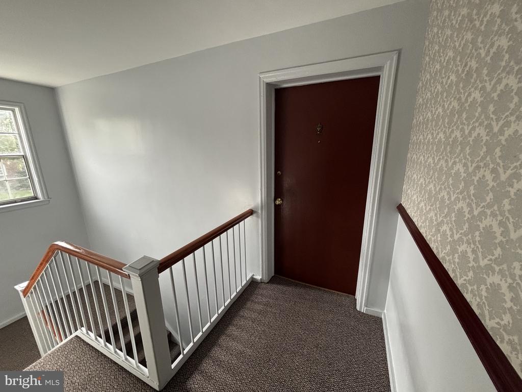 Carpeted stair landing with white railings, patterned wallpaper, and a dark red entry door.