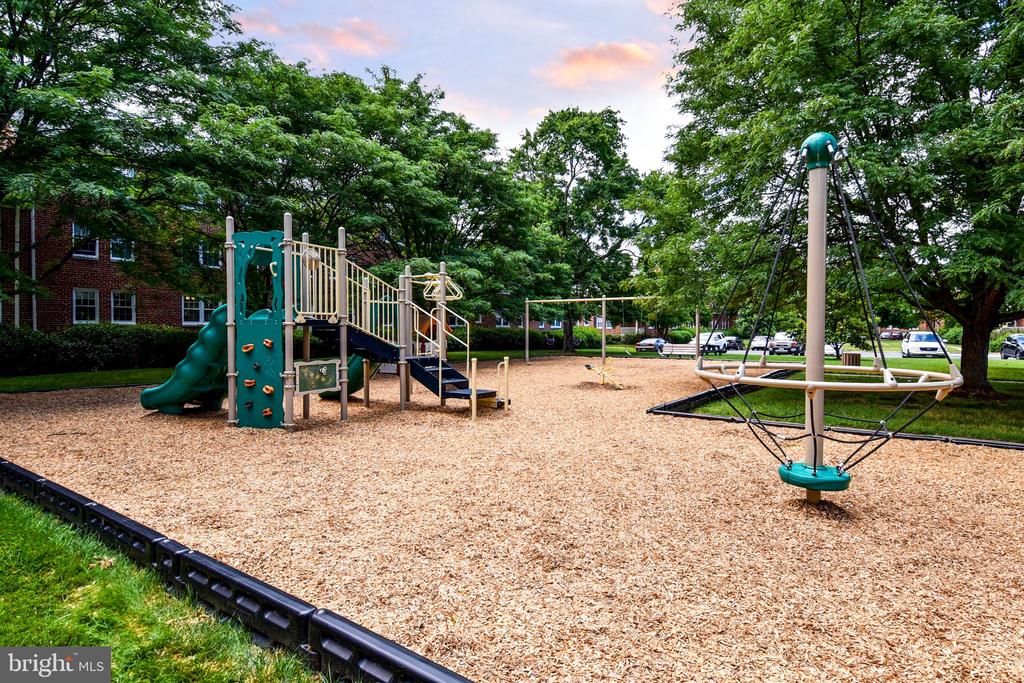 Community playground with a climbing structure and rope spinner beneath mature trees.