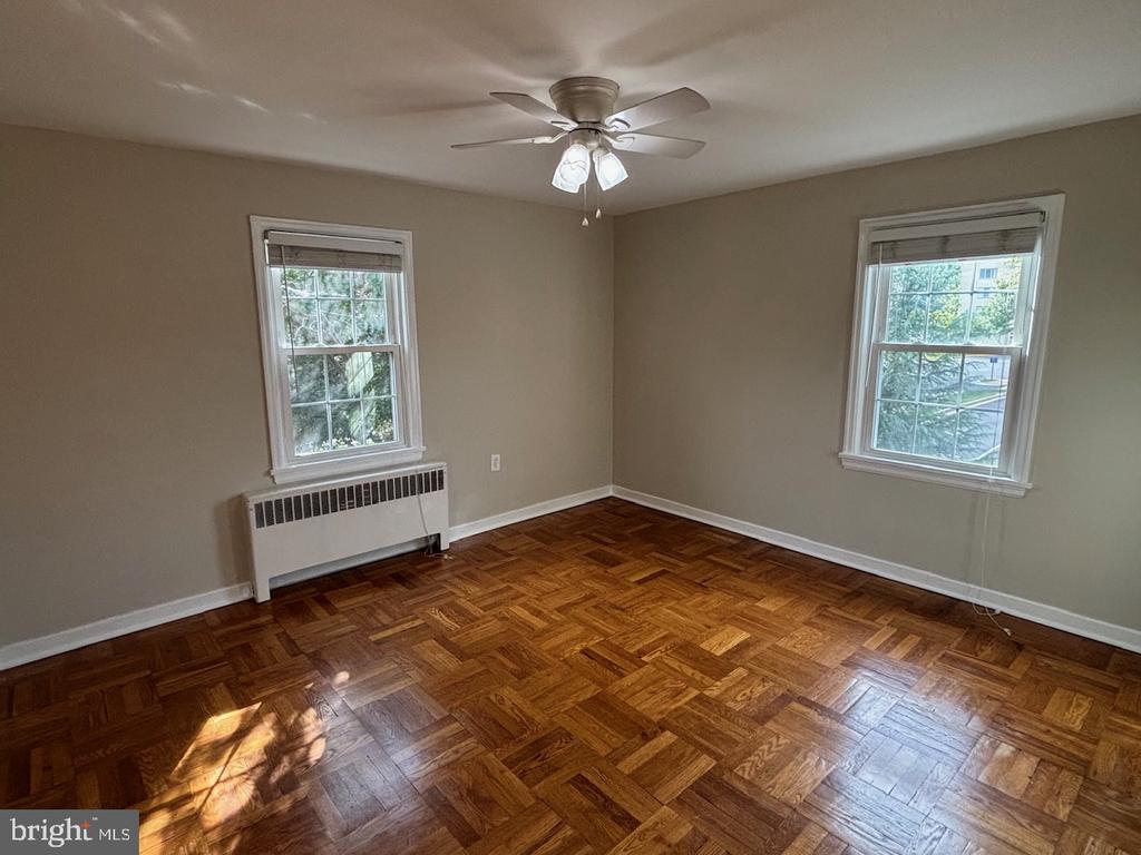 Empty bedroom with parquet floors, two windows, a radiator, and a ceiling fan.