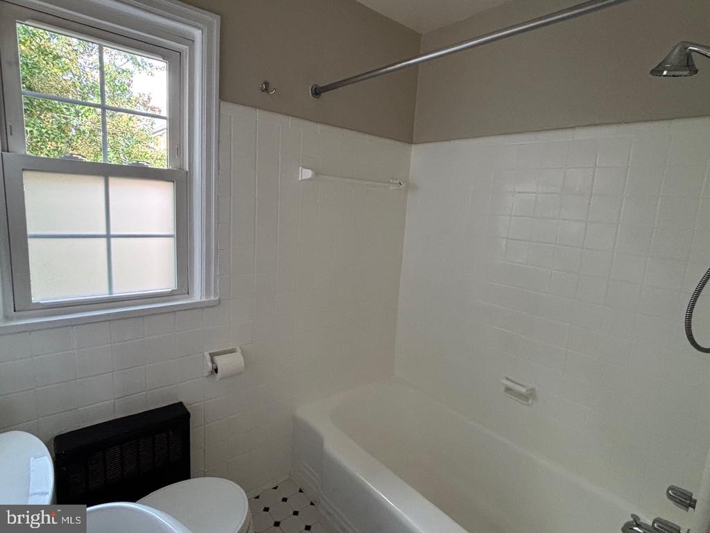 Bathroom with a white tiled tub and shower beside a window and black wall heater.