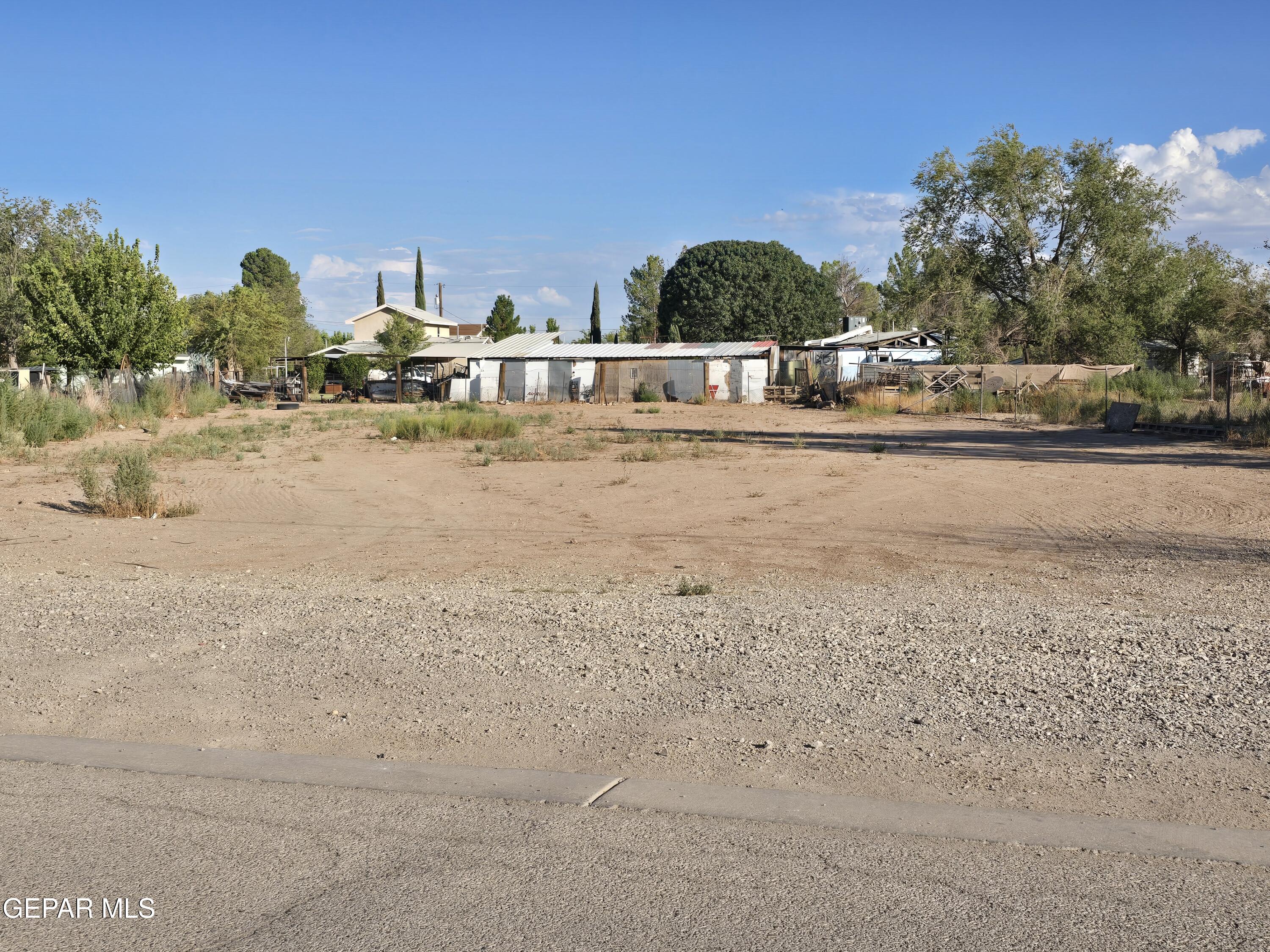 Bare dirt lot with gravel foreground, sparse weeds, fencing, and small outbuildings.