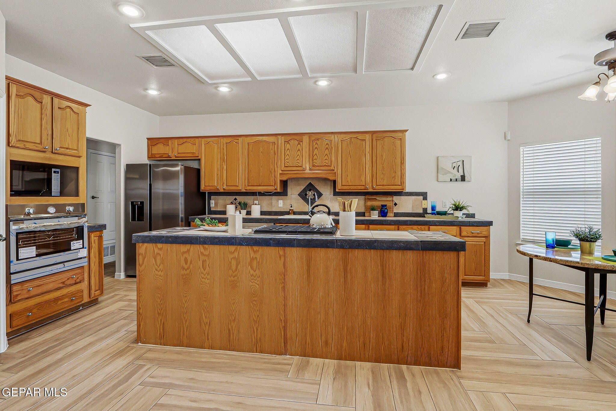Kitchen with honey-colored wood cabinets, a large dark-countertop island, stainless steel appliances, and light tile flooring.