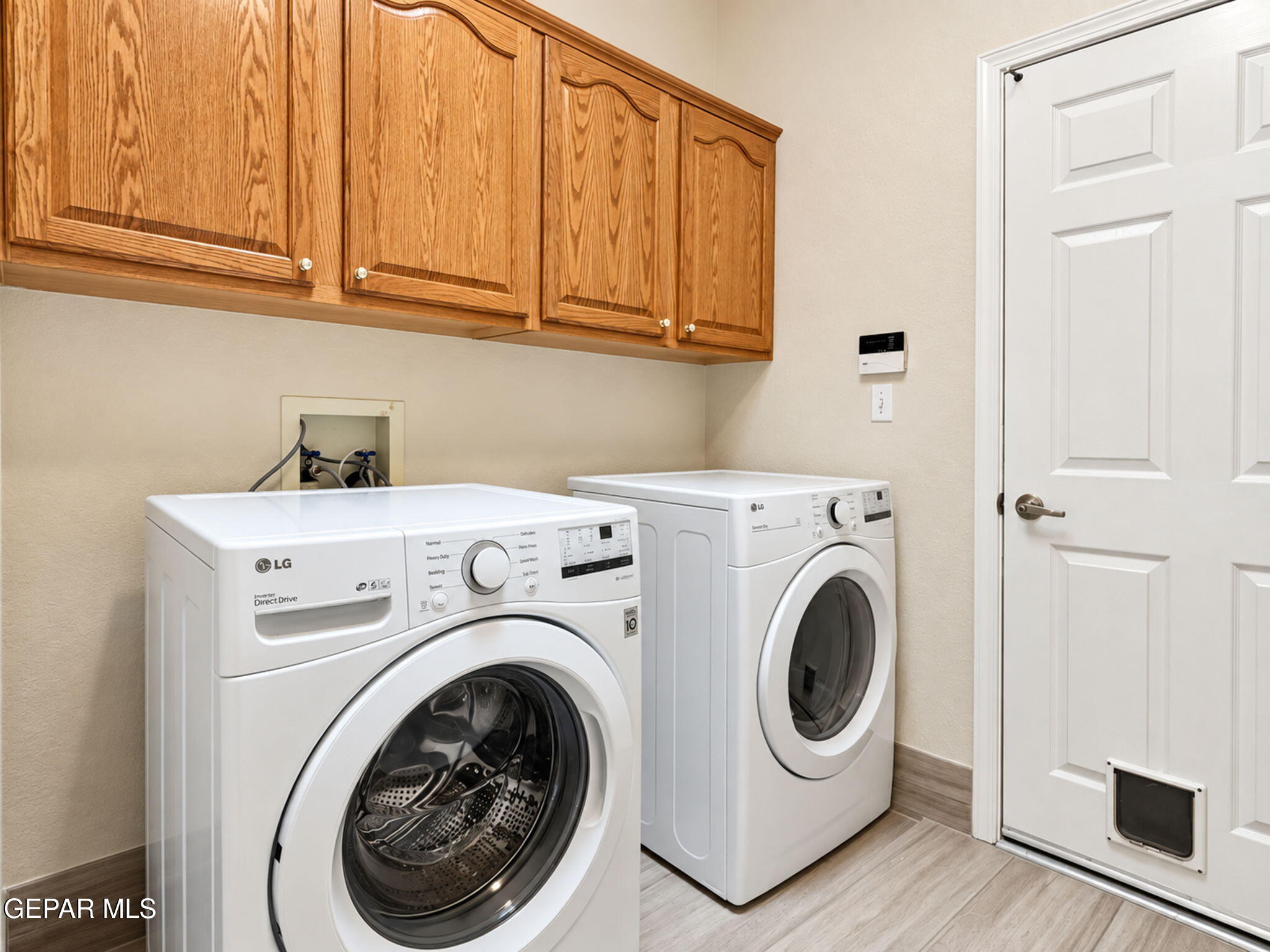 Laundry room with front-loading washer and dryer beneath wooden cabinets, beside a door with a pet flap.