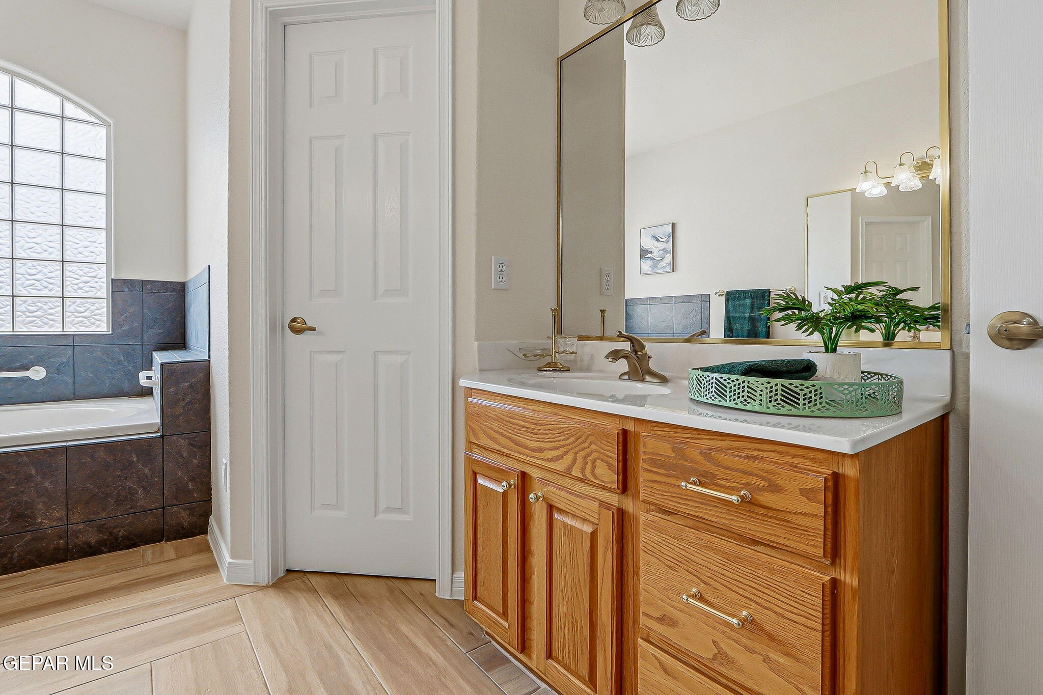 Bathroom with wood vanity and mirror beside a bathtub with dark tile surround and glass-block window.