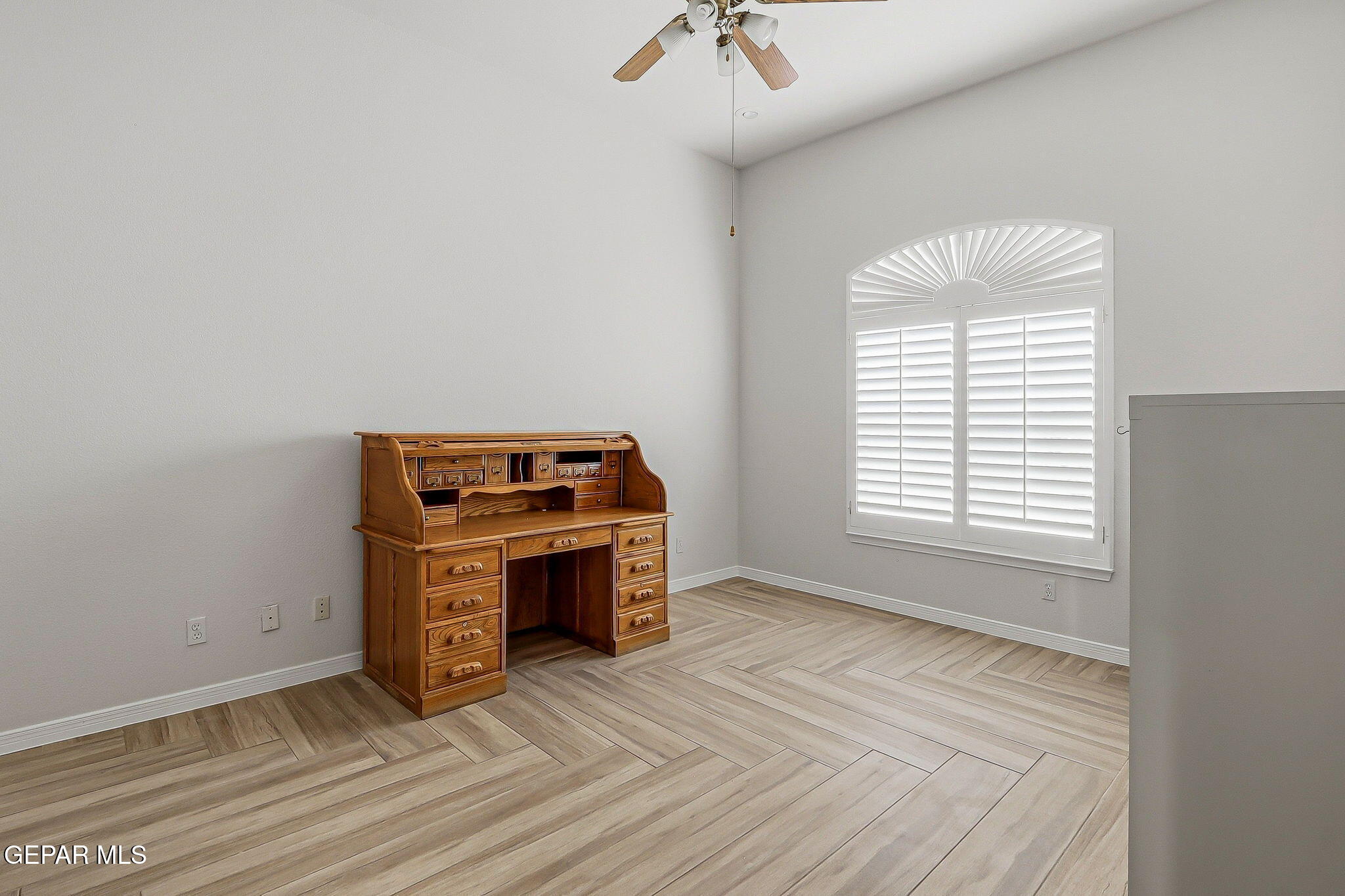 Room with a wooden desk, wood-look tile flooring, and an arched window with plantation shutters.