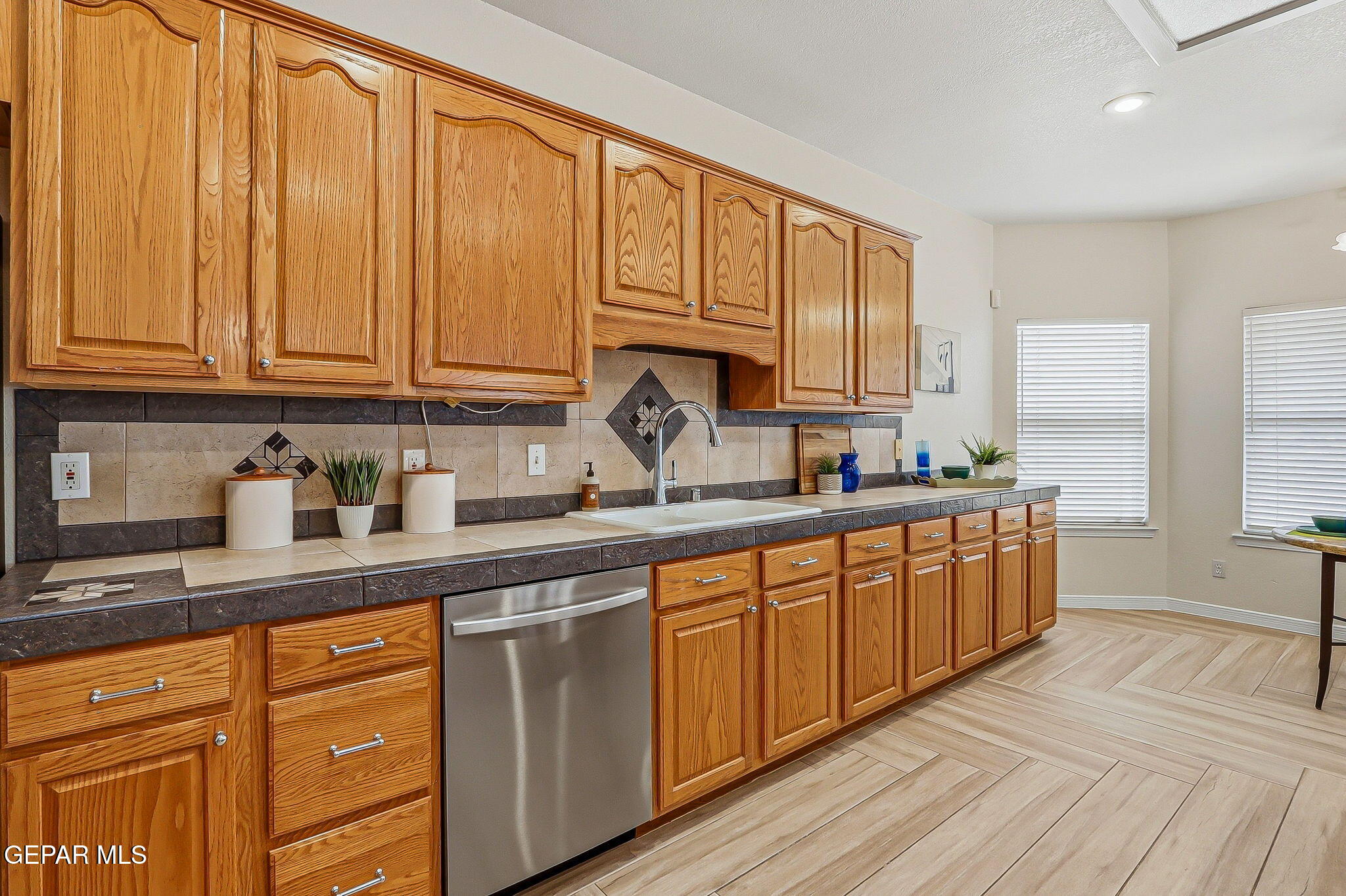 Kitchen with honey-colored wood cabinets, tiled counters, stainless dishwasher, and two windows with blinds.