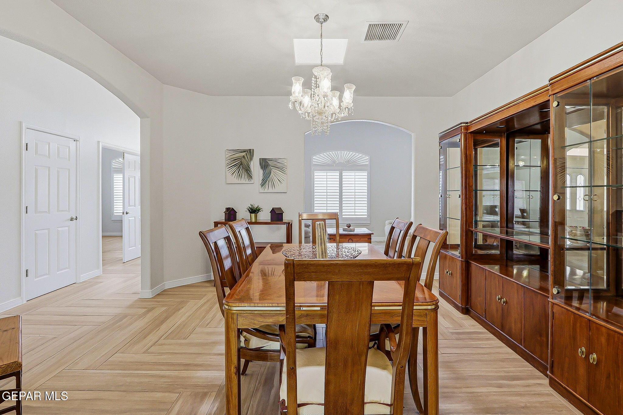 Dining room with a wooden table, glass-front china cabinet, chandelier, and arched shuttered window.