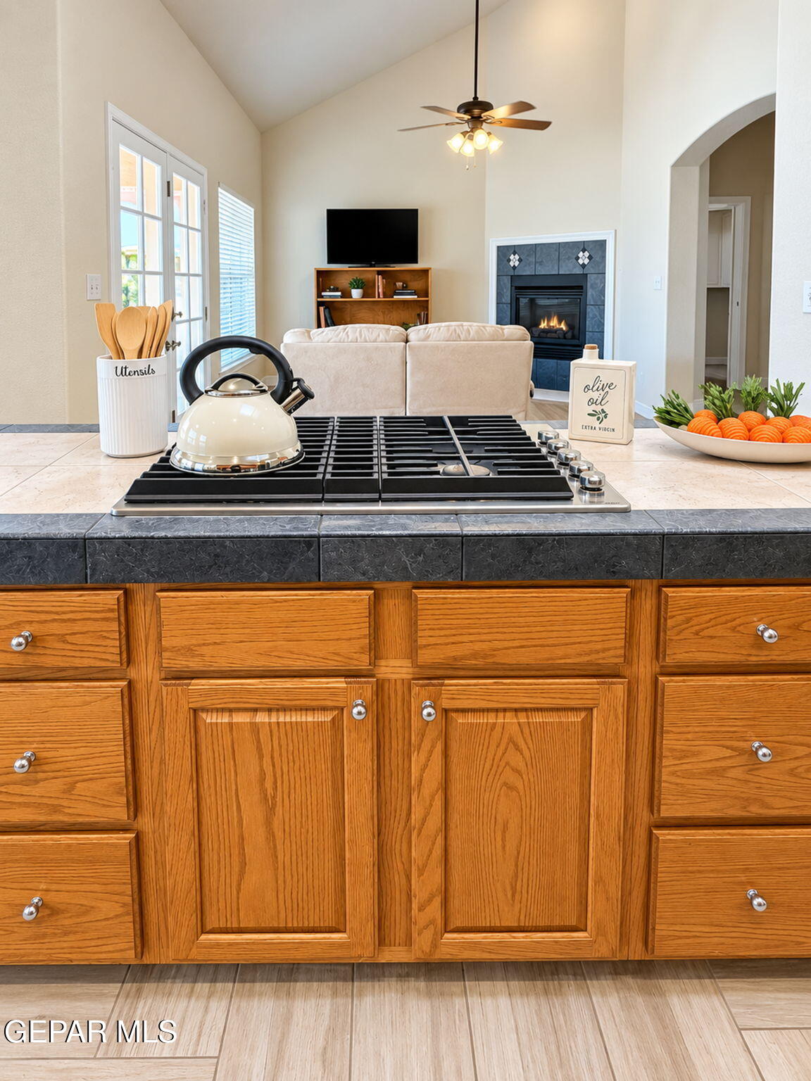 Kitchen with wood cabinetry and gas cooktop opening onto a vaulted living room with a tiled fireplace.