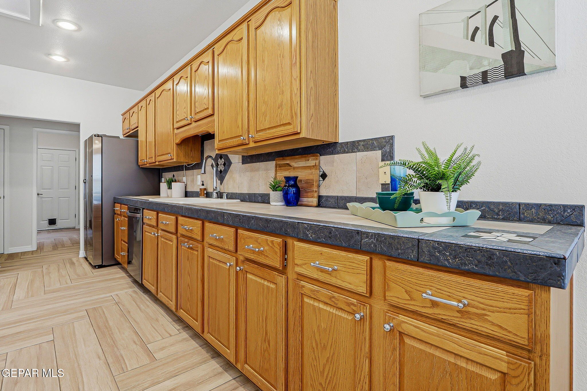 Kitchen with honey-toned wood cabinets, dark counters, stainless steel refrigerator, and decorative plants.