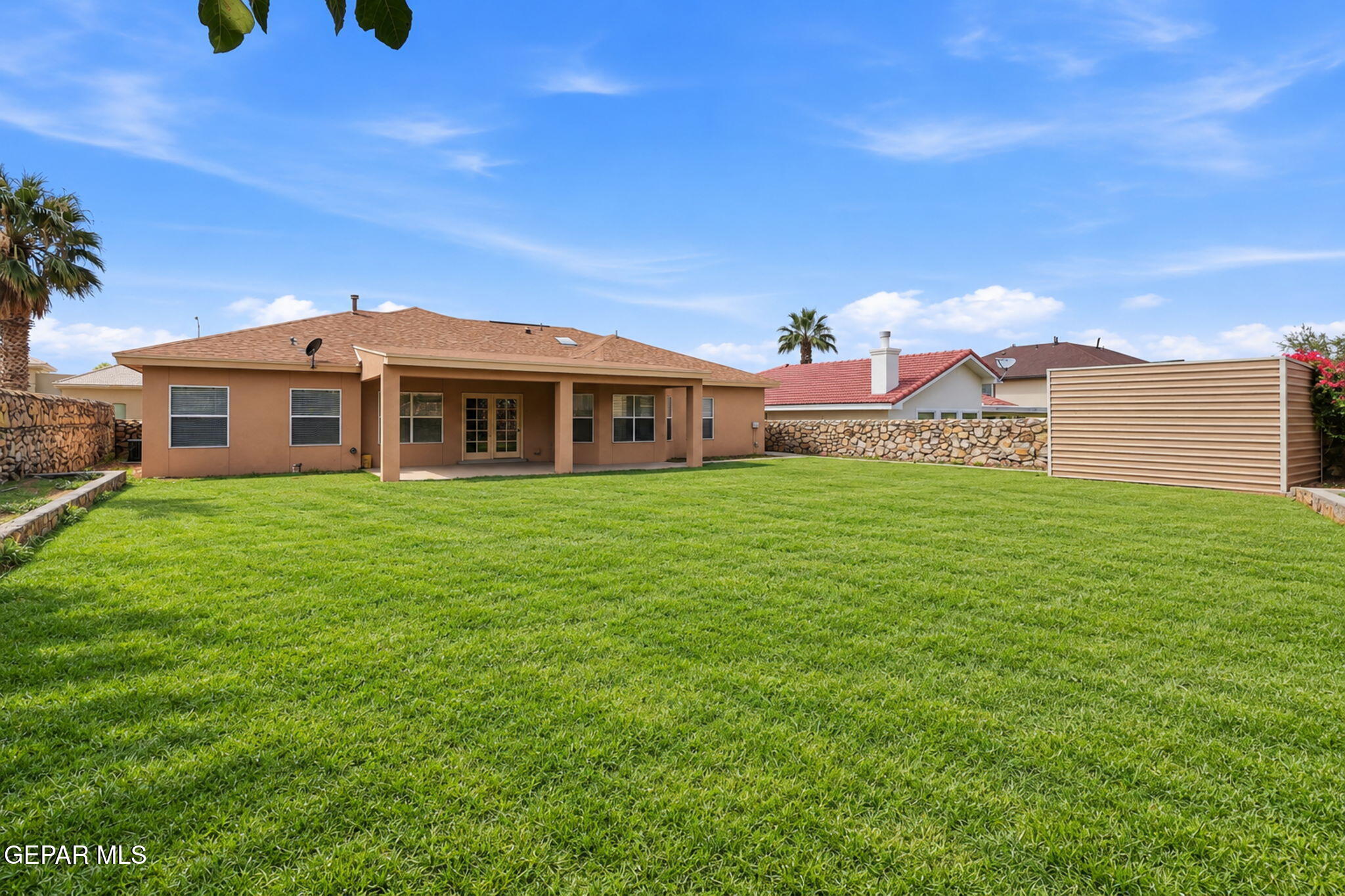 Rear of a tan stucco home with covered patio, broad green lawn, stone wall, and privacy screen.