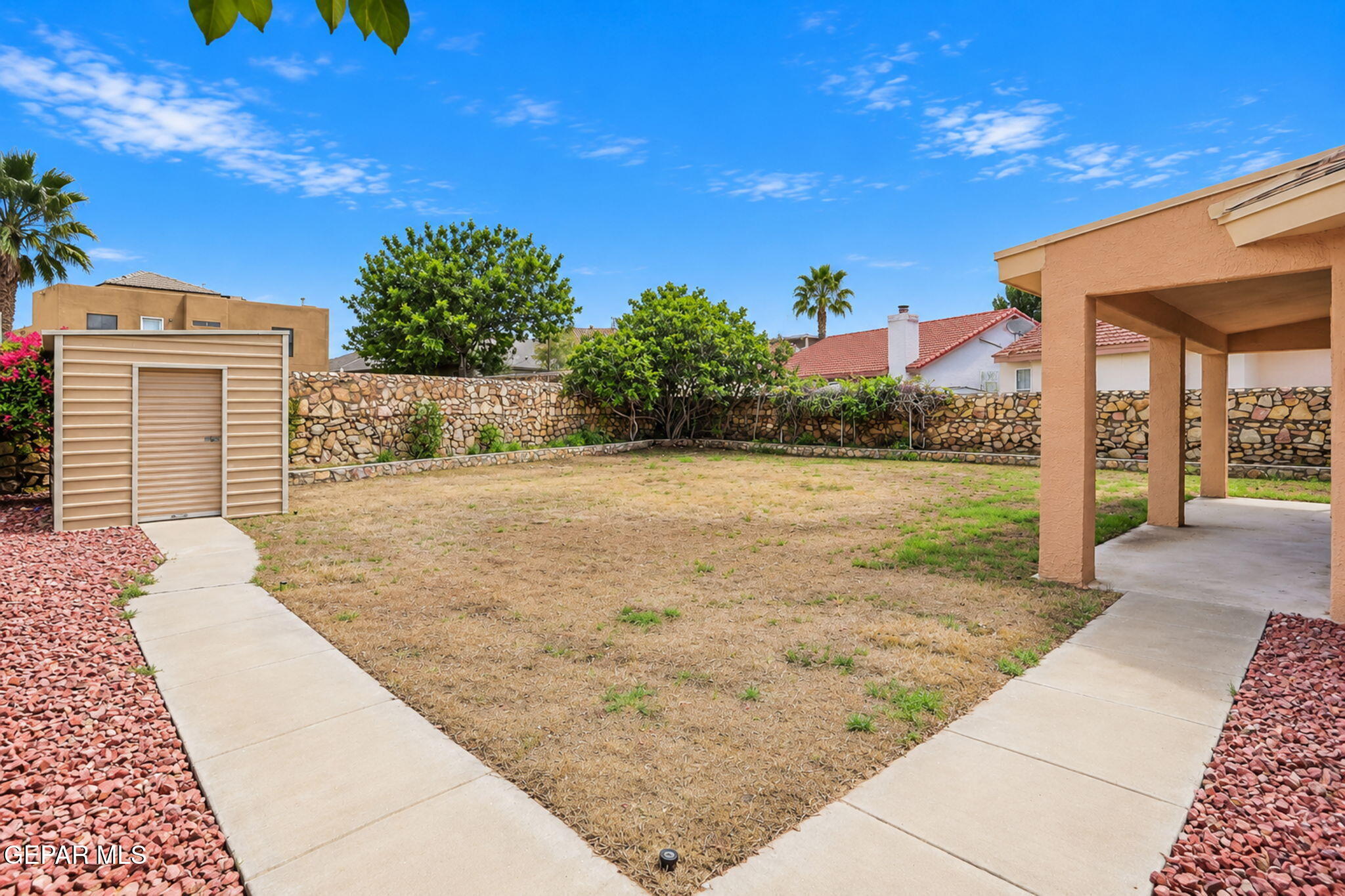 Backyard with a covered patio, dry lawn, storage shed, and stone boundary wall.