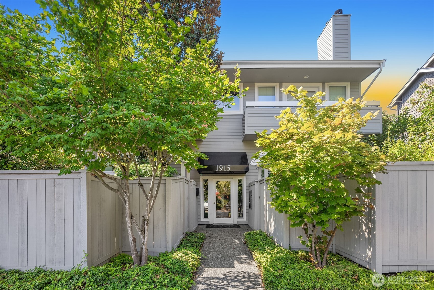Gray two-story home with an upper balcony, white fencing, and a tree-lined walkway to the entry.