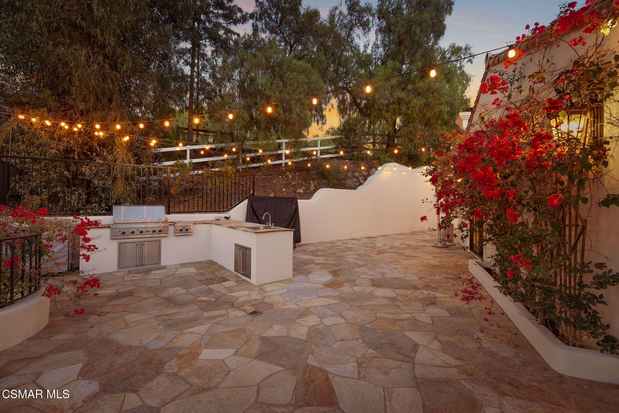 Stone patio with an outdoor kitchen, red flowering vines, and glowing string lights at dusk.