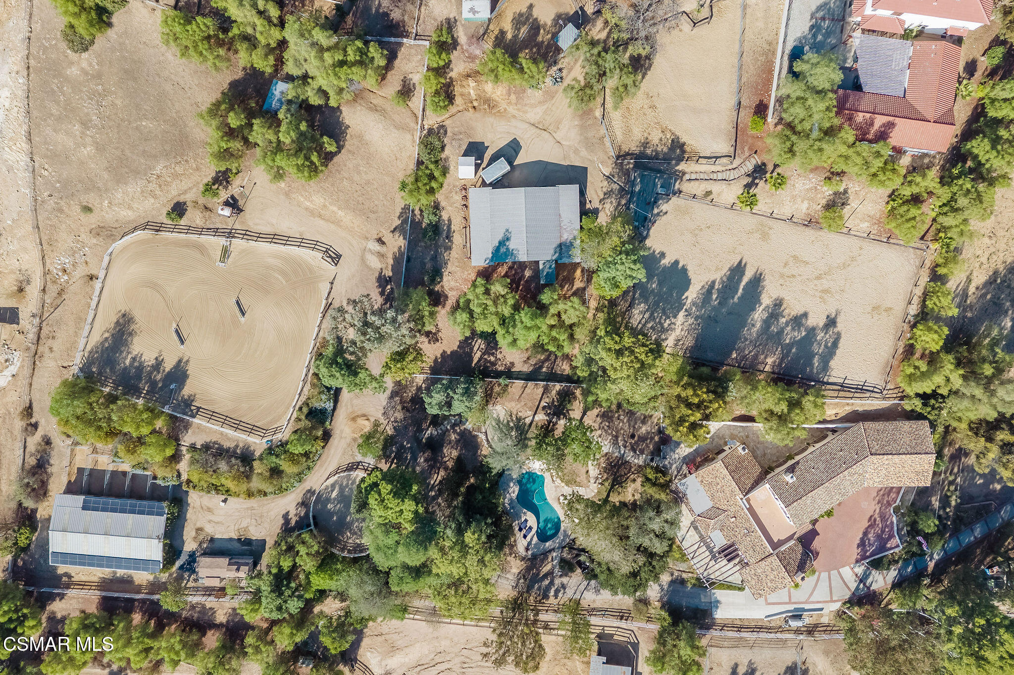 Aerial view of a wooded property with a tiled-roof home, pool, riding arena, and fenced paddocks.