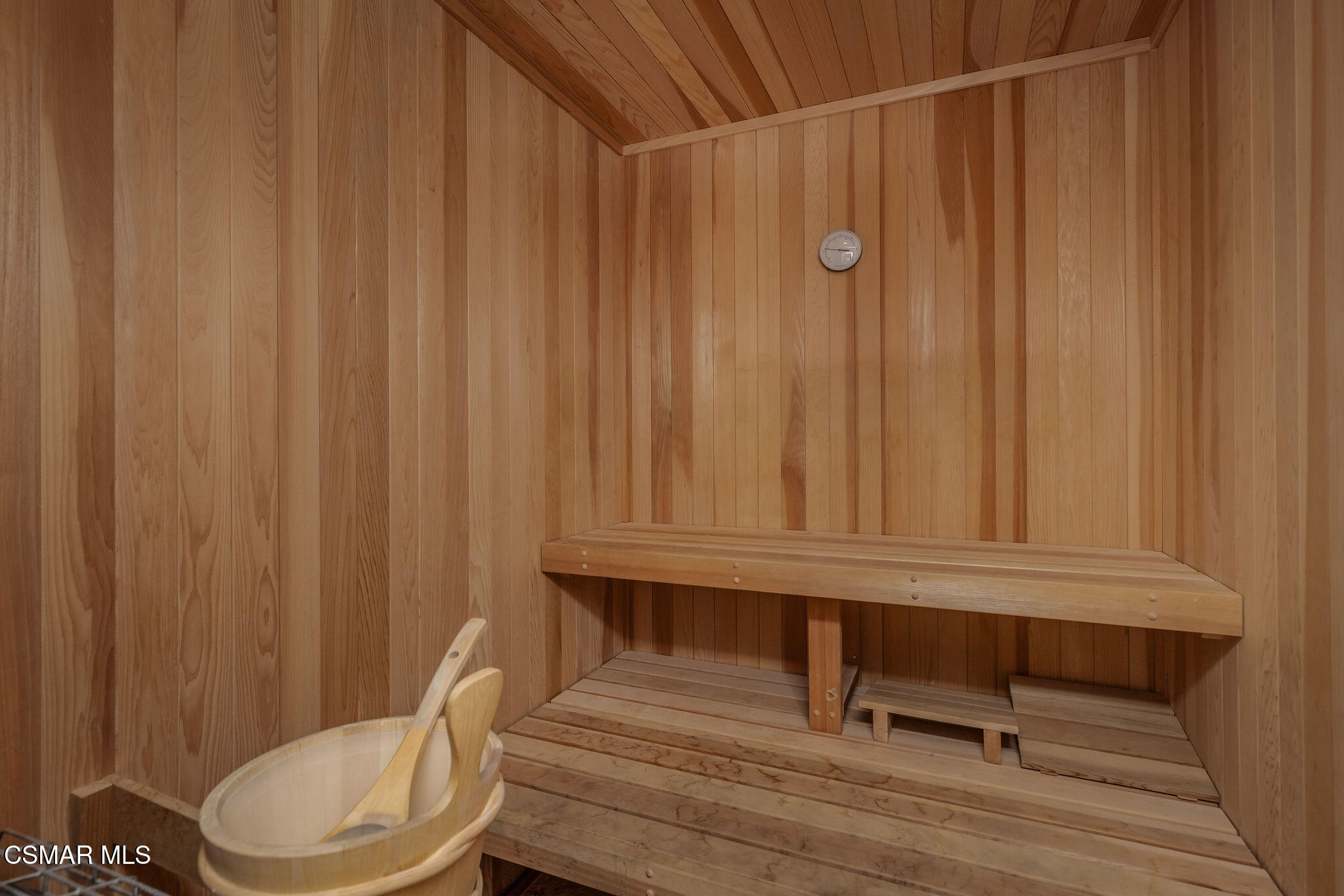 Cedar-paneled sauna with tiered benches, a wall thermometer, and a bucket with ladle.