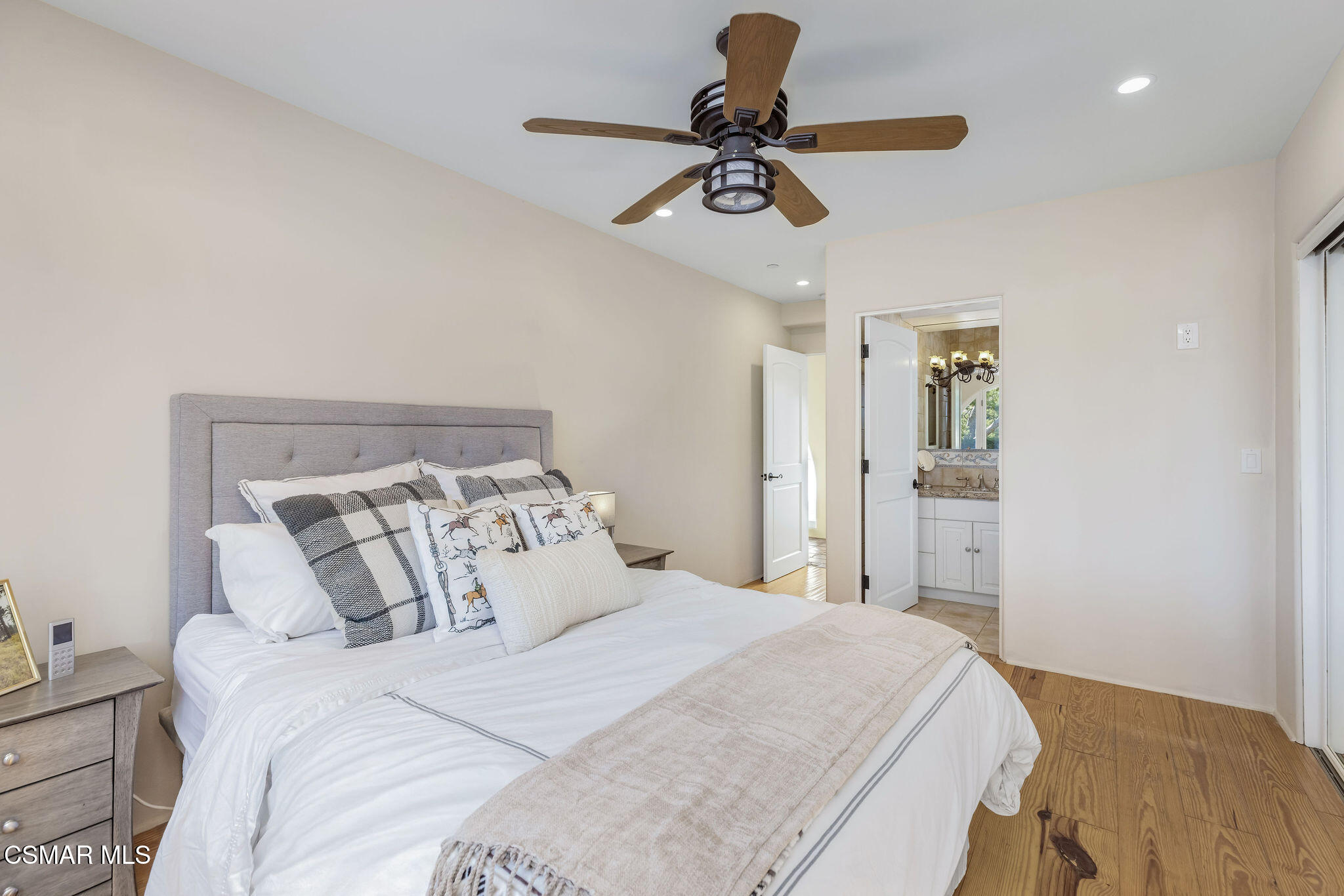 Staged bedroom with a white bed, wood ceiling fan, hardwood floors, and an open en-suite bathroom doorway.