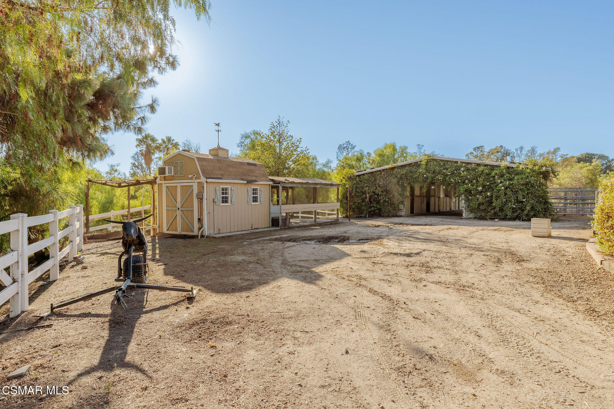 Equestrian yard with a tan barn, covered stalls, and white fencing.