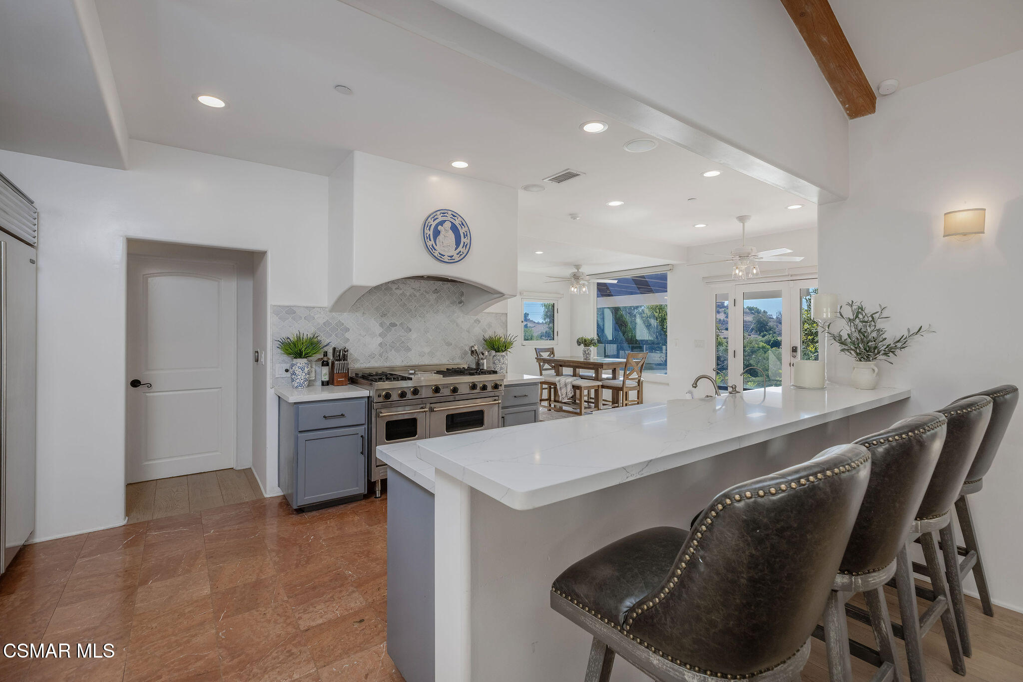 Open kitchen with a white waterfall island, dark bar stools, and a dining area overlooking green hills.