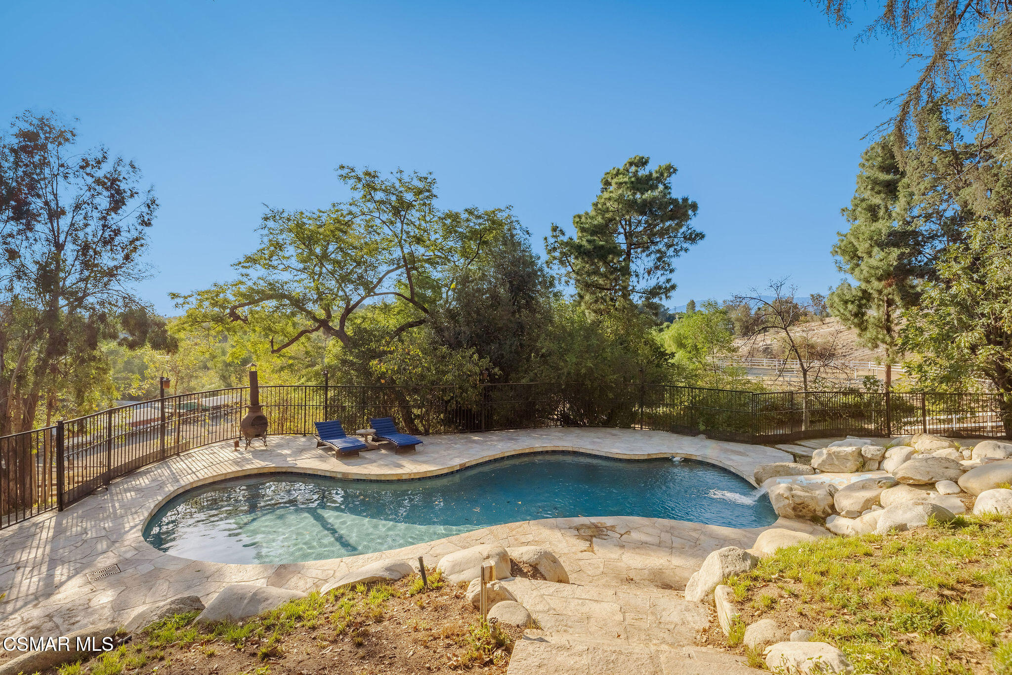 Backyard swimming pool with a stone patio, blue lounge chairs, and mature trees.
