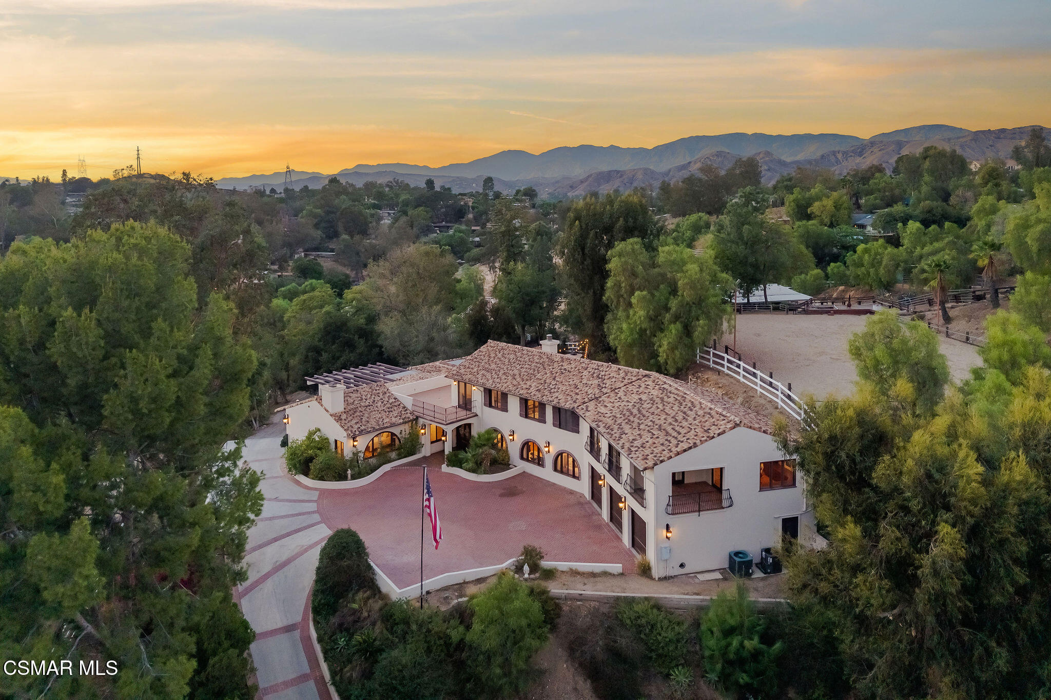 Aerial view of a Spanish-style home with a circular brick driveway, fenced paddock, and mountain backdrop at dusk.