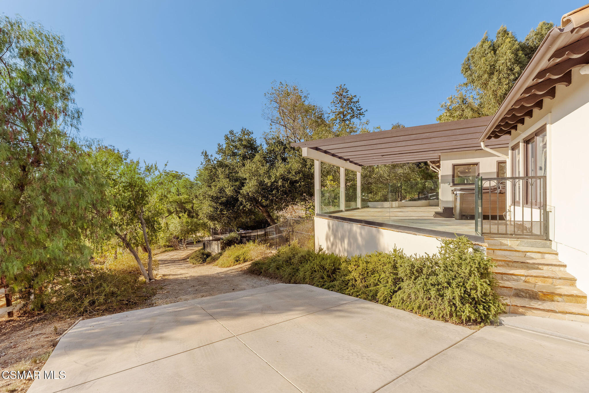 Home patio with a covered raised deck, glass railings, hot tub, and mature trees.