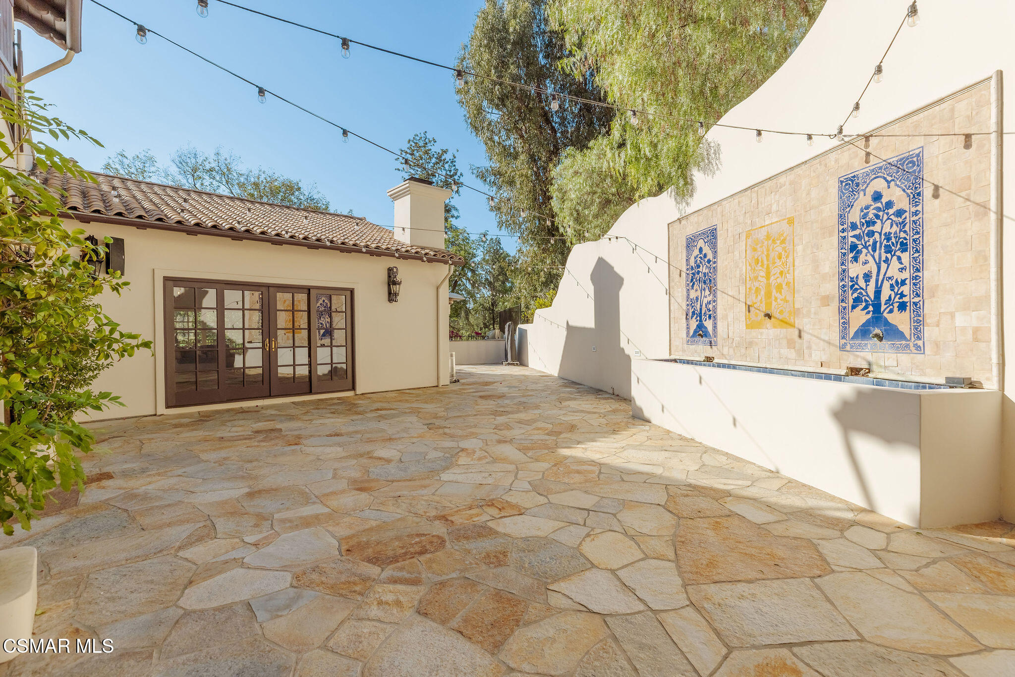 Stone patio with French doors, string lights, and a tiled wall fountain with blue and yellow panels.