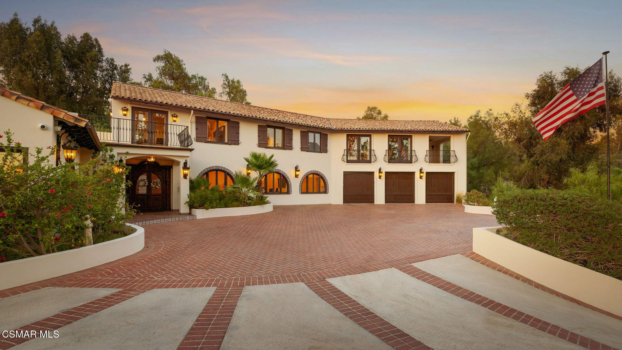 Two-story Spanish-style home with arched windows, three garage doors, and a broad brick driveway at dusk.