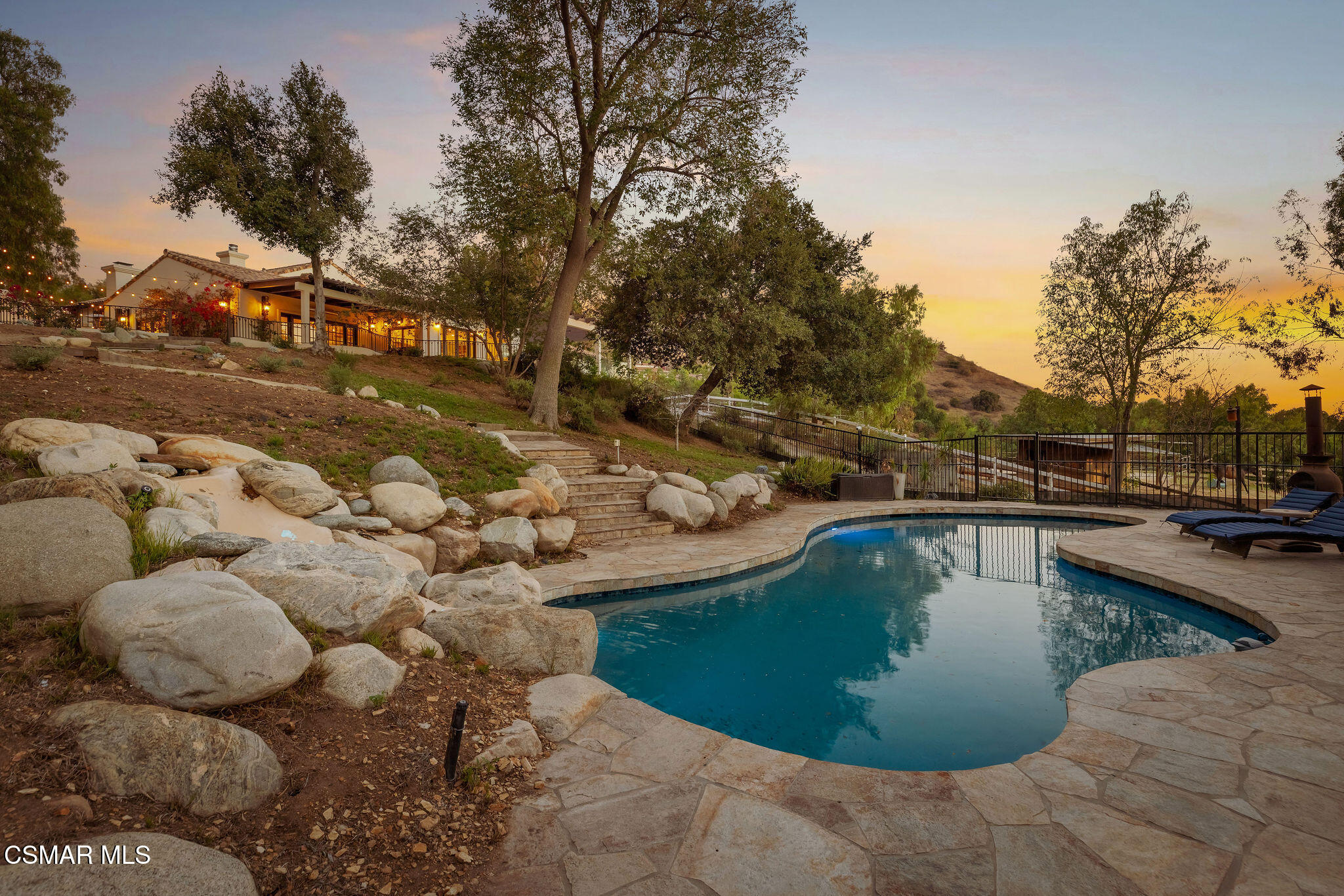 Hillside home and curving pool surrounded by boulders and mature trees at sunset.