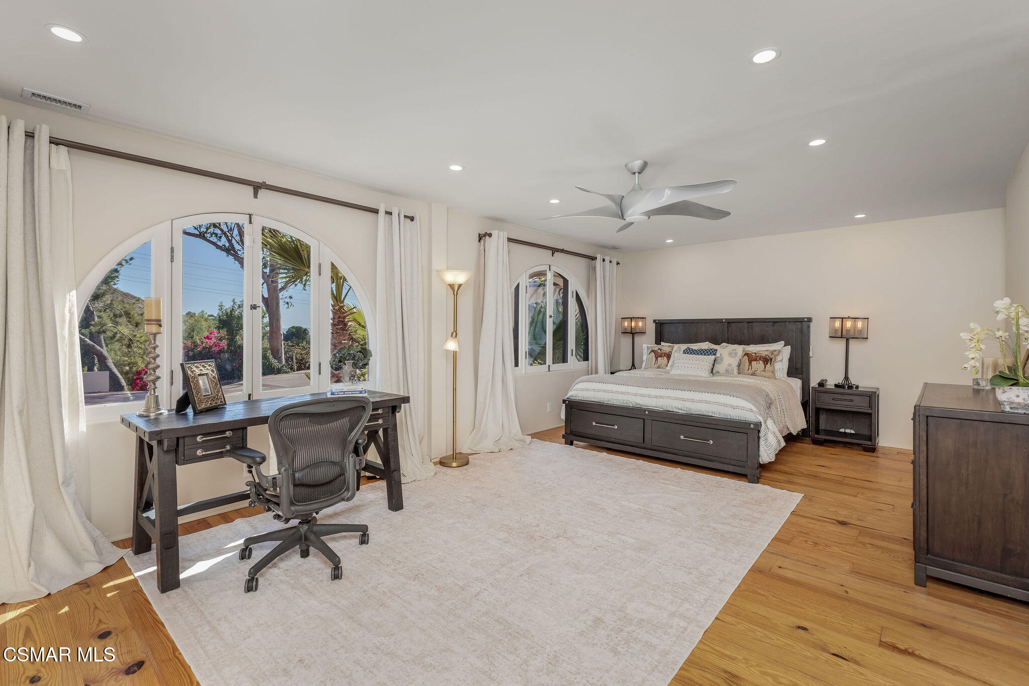 Furnished bedroom with arched windows, a dark wood bed, desk, and hardwood floors.