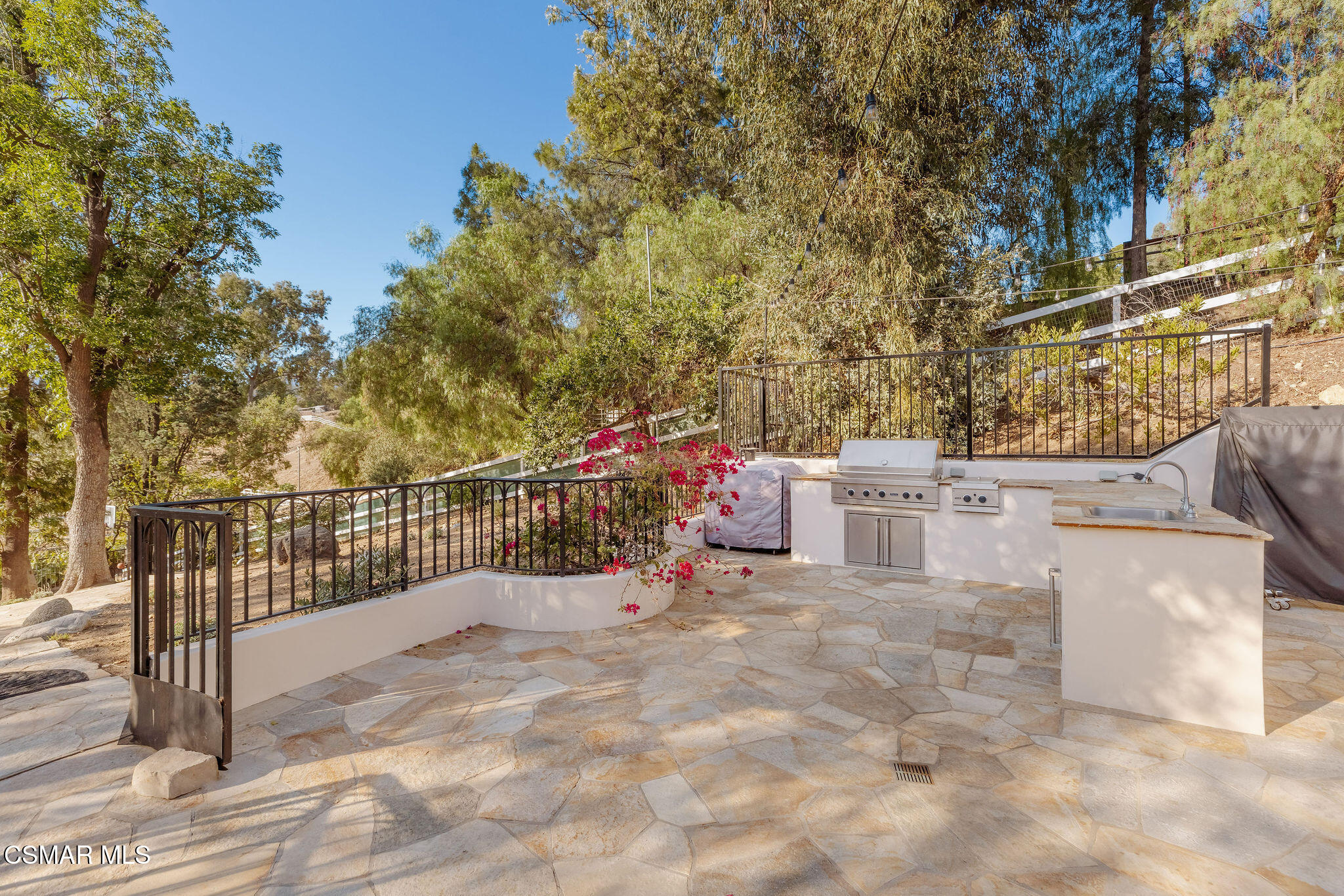 Stone patio with built-in grill and sink, bordered by black railings and flowering bougainvillea.