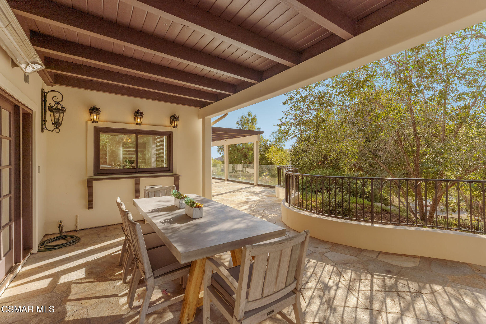 Covered patio with a dining table and chairs, stone flooring, and a curved railing beside leafy trees.