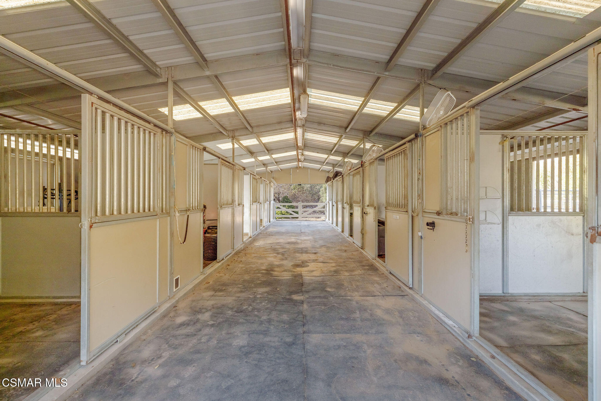 Stable aisle lined with horse stalls beneath a high metal roof with skylights.