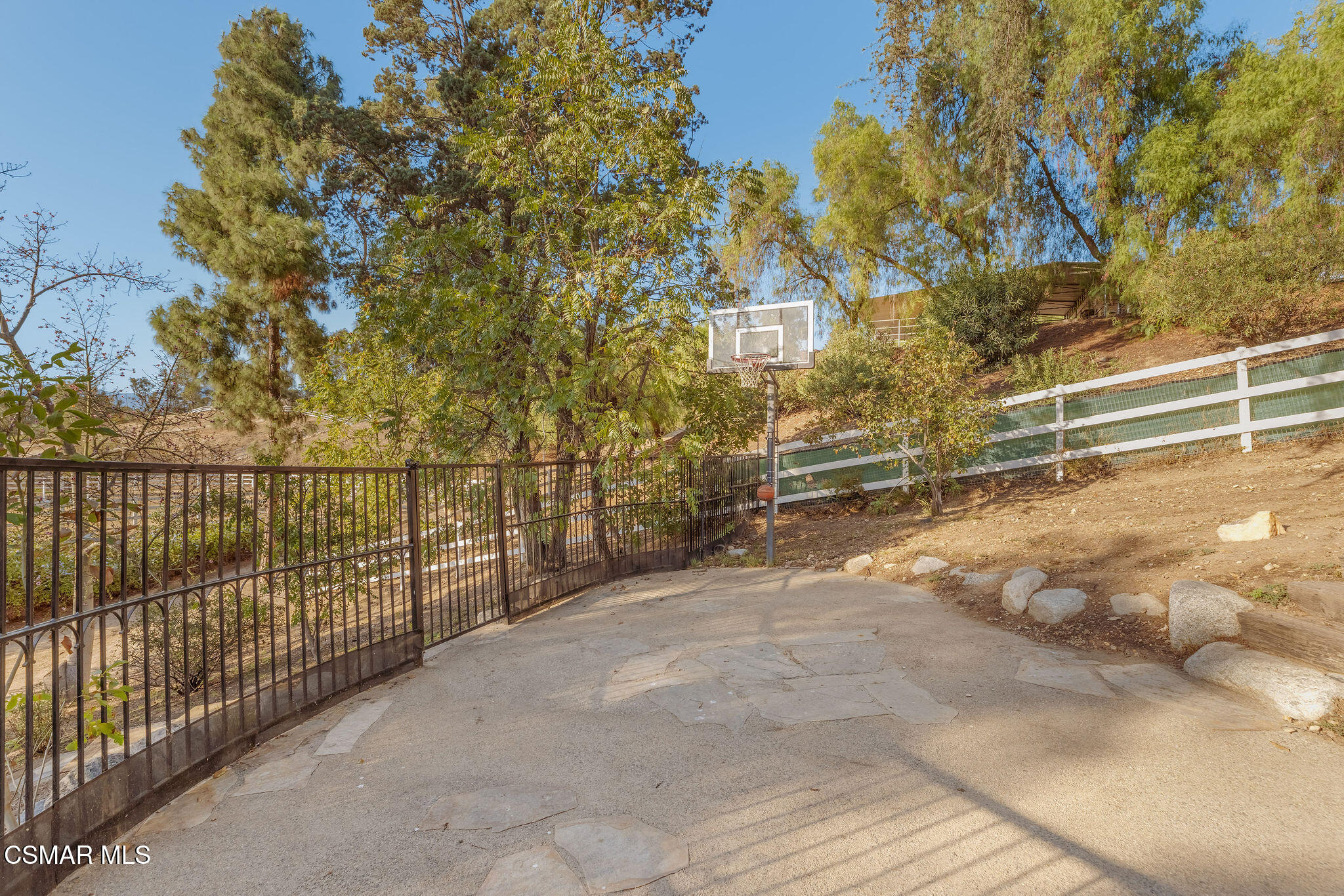 Outdoor basketball court with a hoop, black metal gate, and wooded hillside.