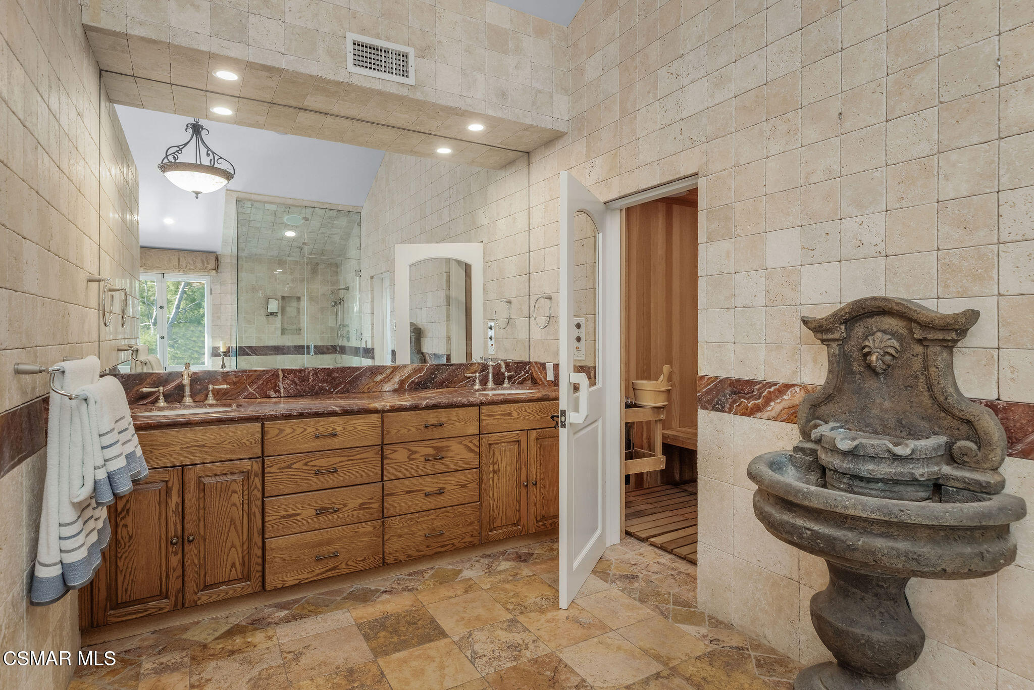 Tiled bathroom with a wooden vanity, glass shower, open sauna doorway, and carved stone fountain.