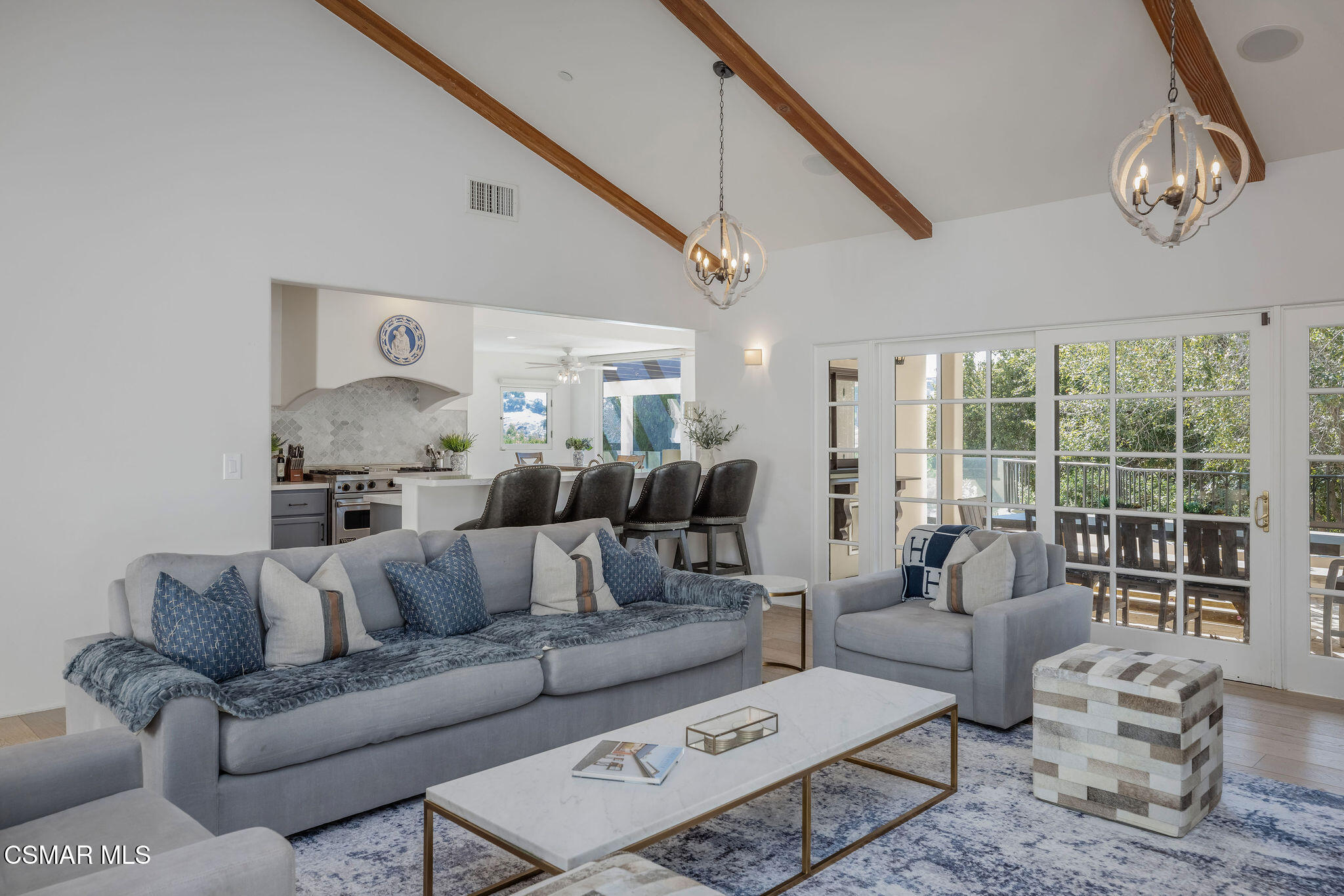 Living room with gray seating and vaulted wood-beam ceiling, opening to a kitchen and glass doors.