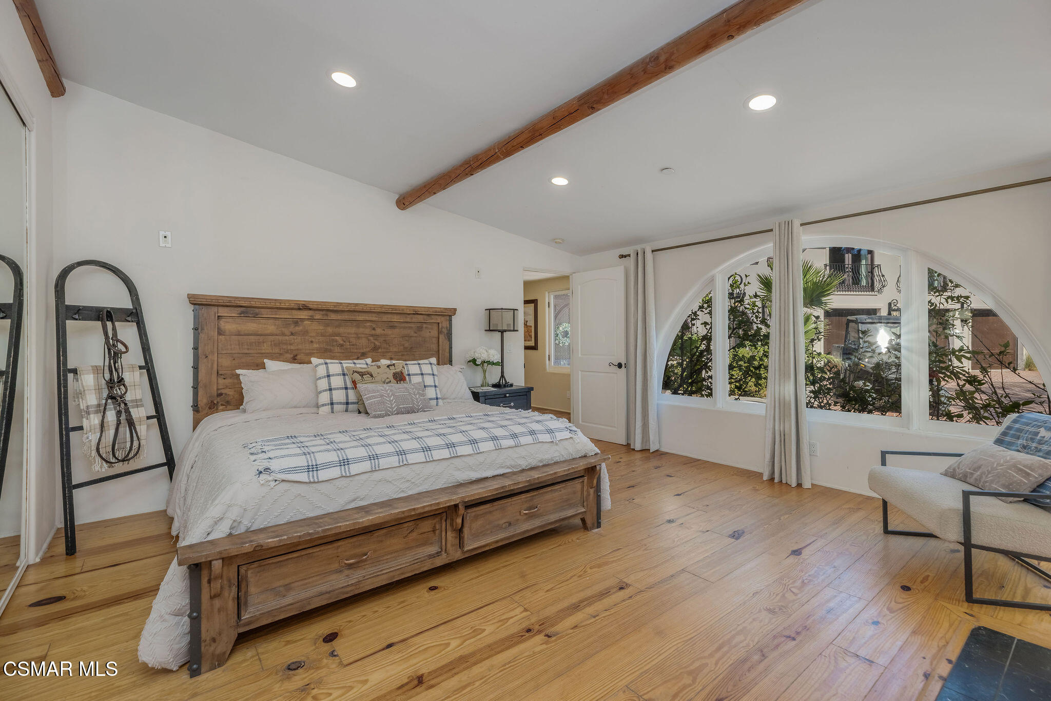 Bedroom with a rustic wooden bed, exposed ceiling beams, wood floors, and arched windows.