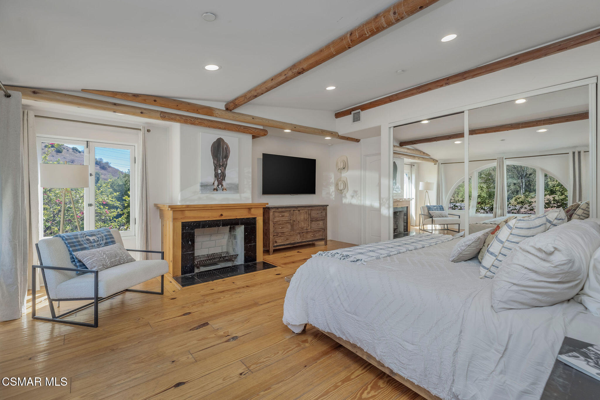 Bedroom with exposed wood beams, hardwood floors, and a fireplace beside a large white bed.