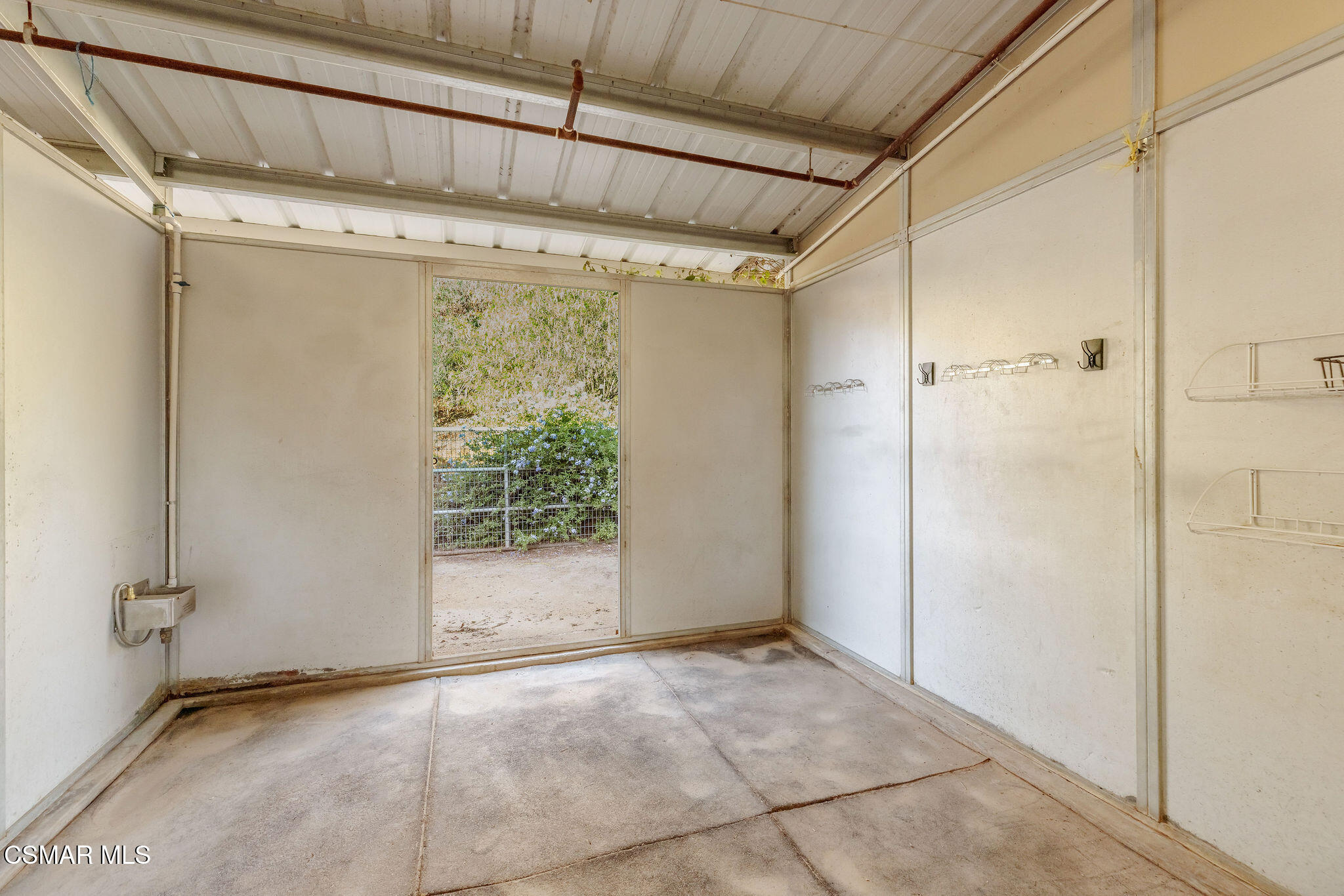 Empty covered garage with concrete floor, wall-mounted racks, and an open doorway to a shrub-lined yard.