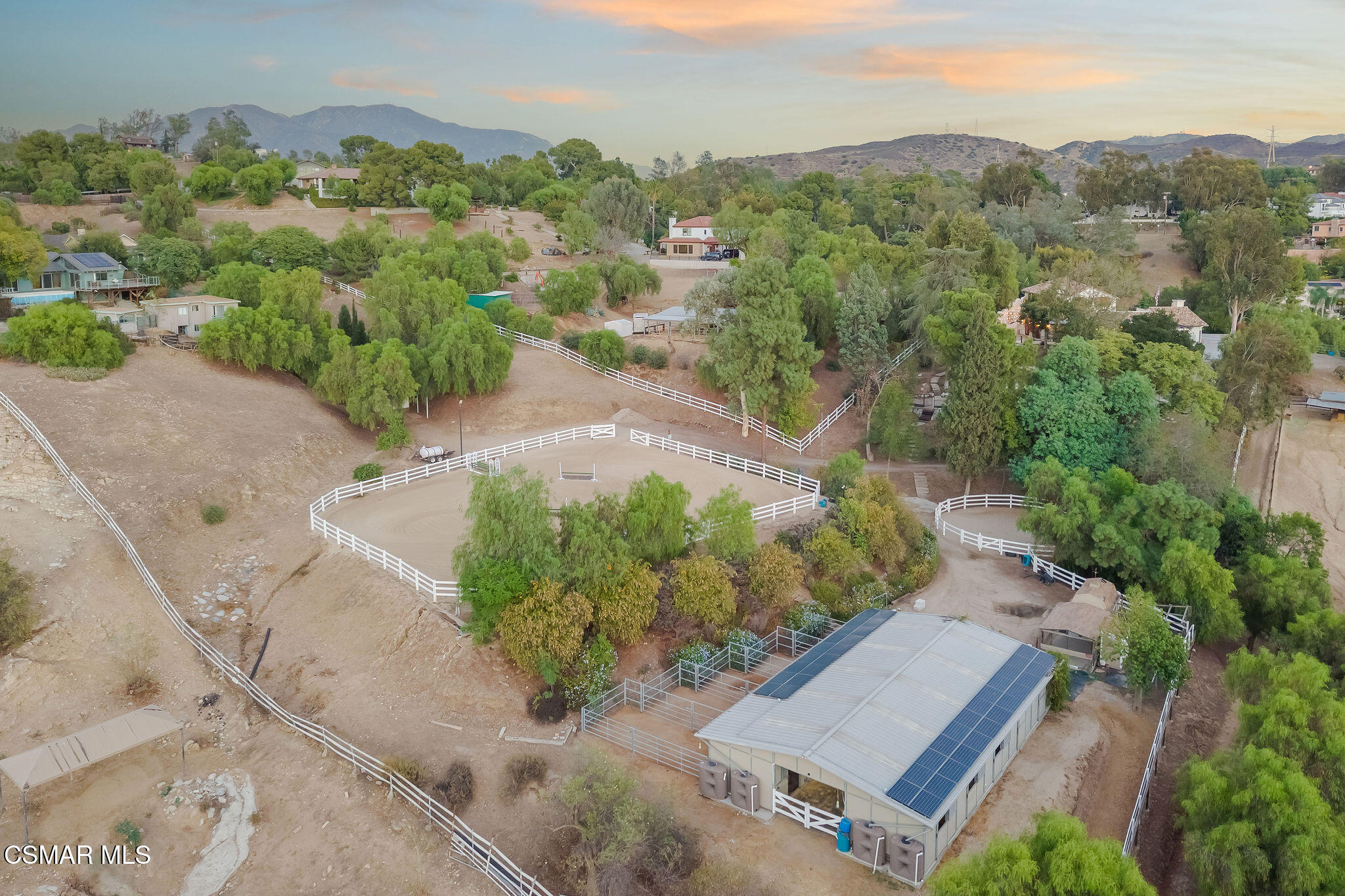 Aerial view of a fenced equestrian property with riding arenas and a solar-panelled outbuilding, backed by mountains.