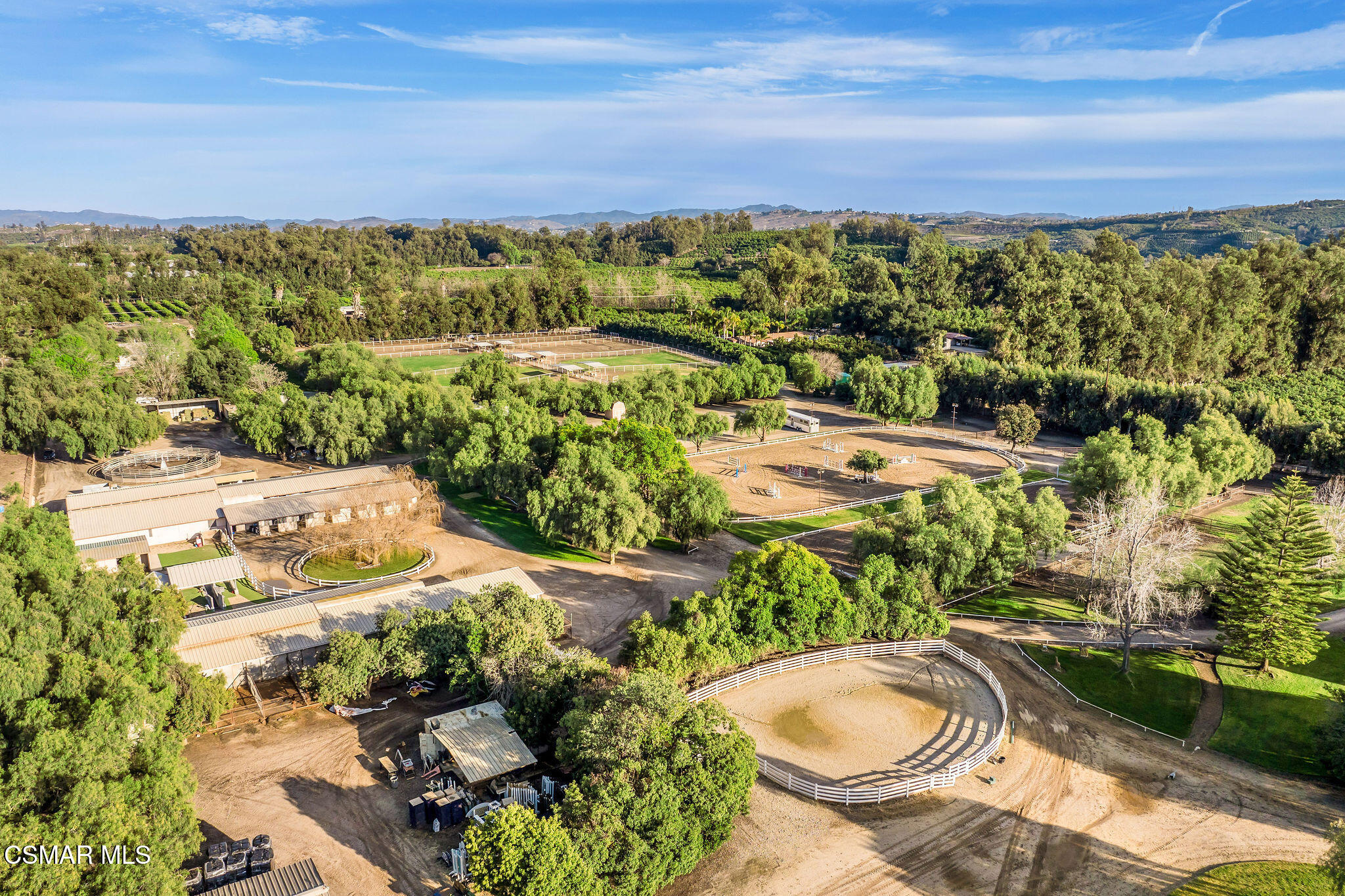 Aerial view of an equestrian property with fenced arenas, round pens, and stable buildings among mature trees.