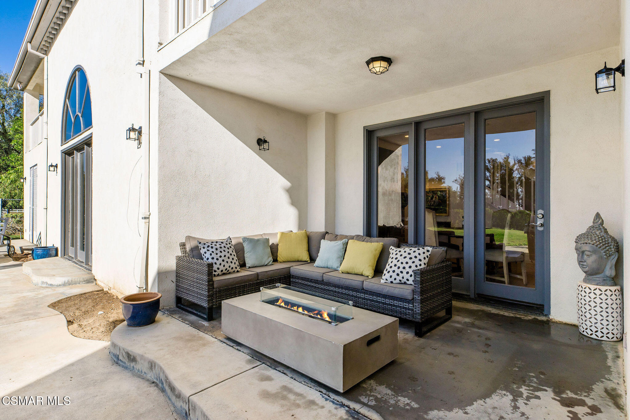 Covered patio with a wicker sectional, colorful cushions, and a lit fire table beside glass doors.