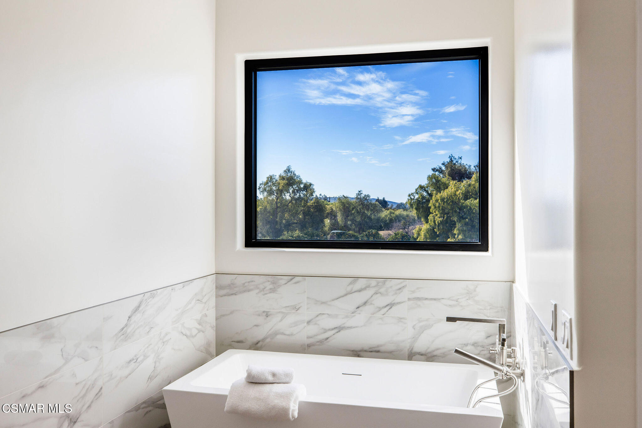 Modern bathroom with a freestanding tub beneath a large window overlooking trees.