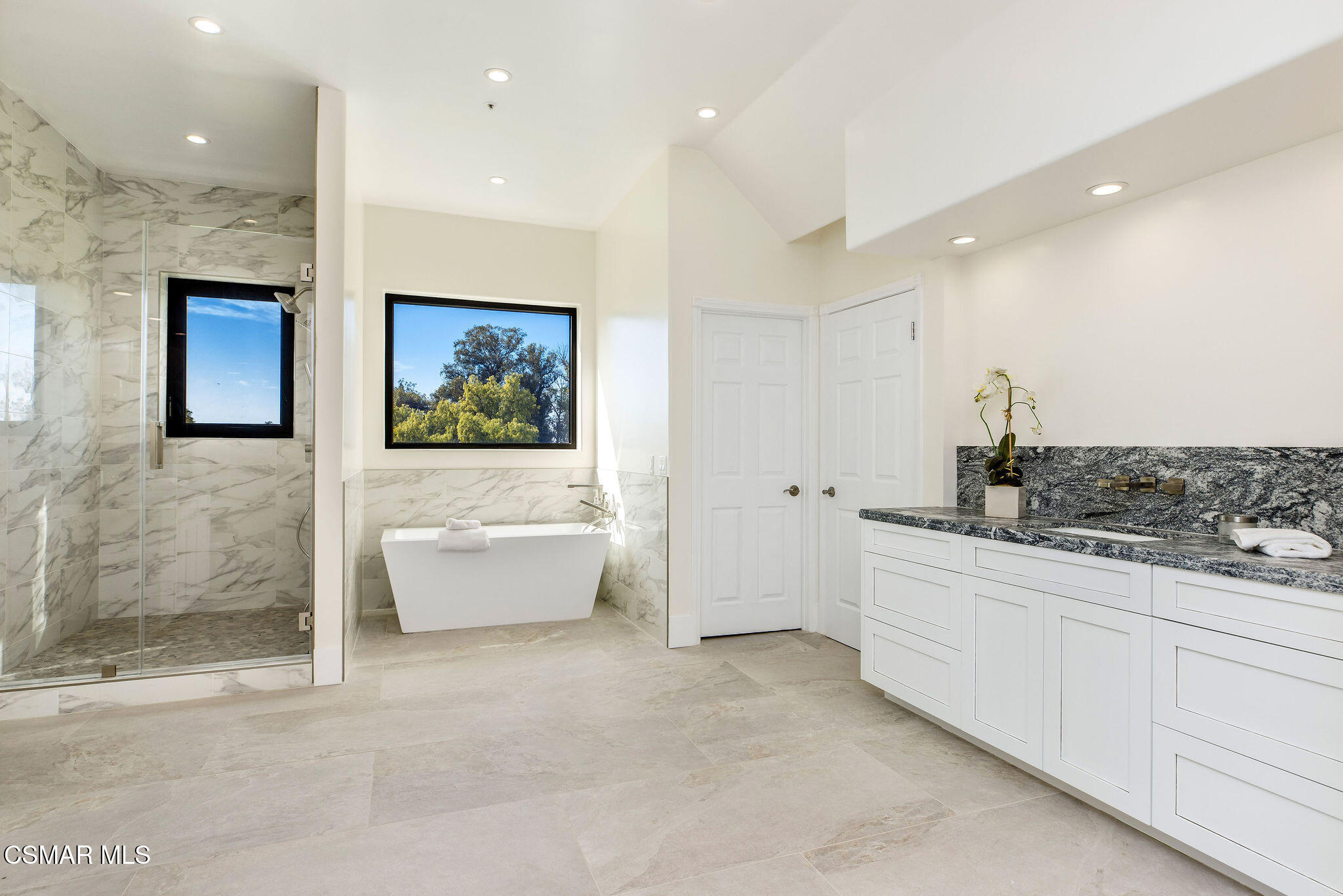 Bright bathroom with a marble shower, freestanding tub beneath a window, and white vanity with dark stone counter.