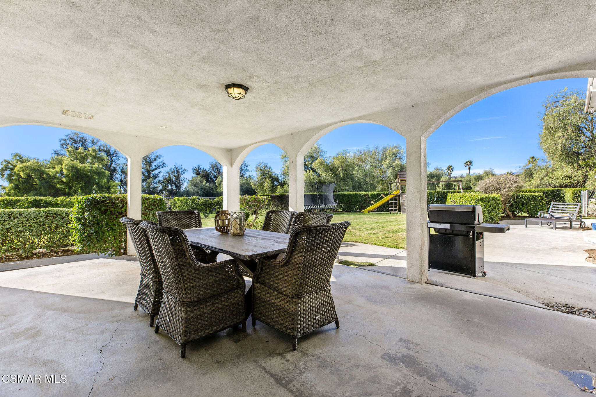 Covered patio with arched openings, wicker dining set, barbecue, and a fenced backyard with play equipment.