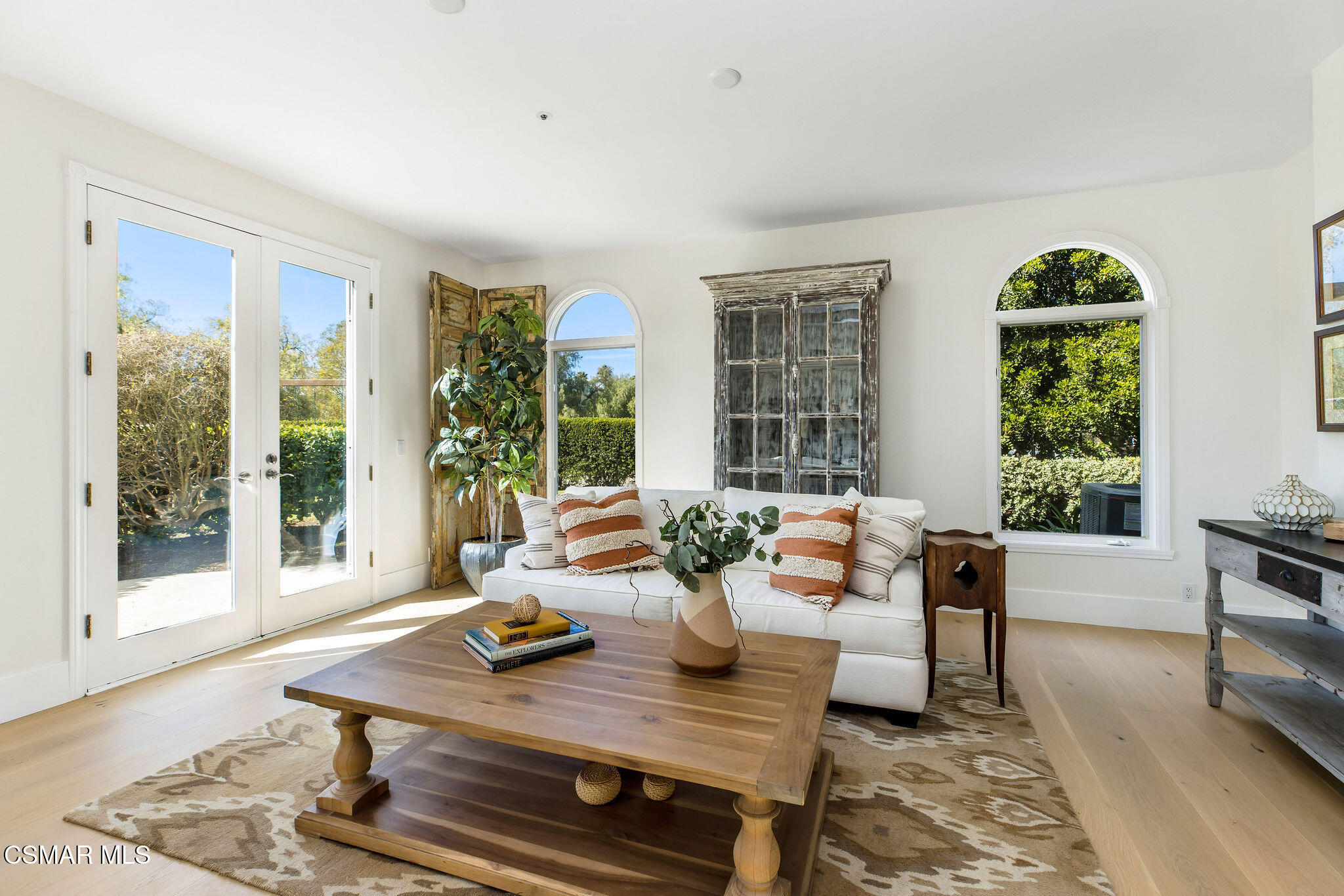 Furnished living room with a white sofa, large wooden coffee table, and arched windows overlooking greenery.