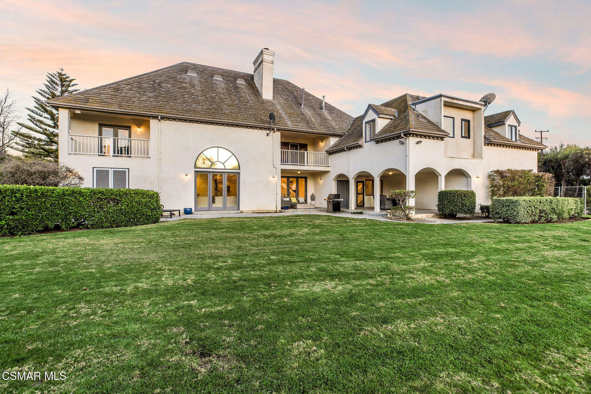 Large cream stucco home with balconies and arched patio overlooking a broad lawn at golden hour.